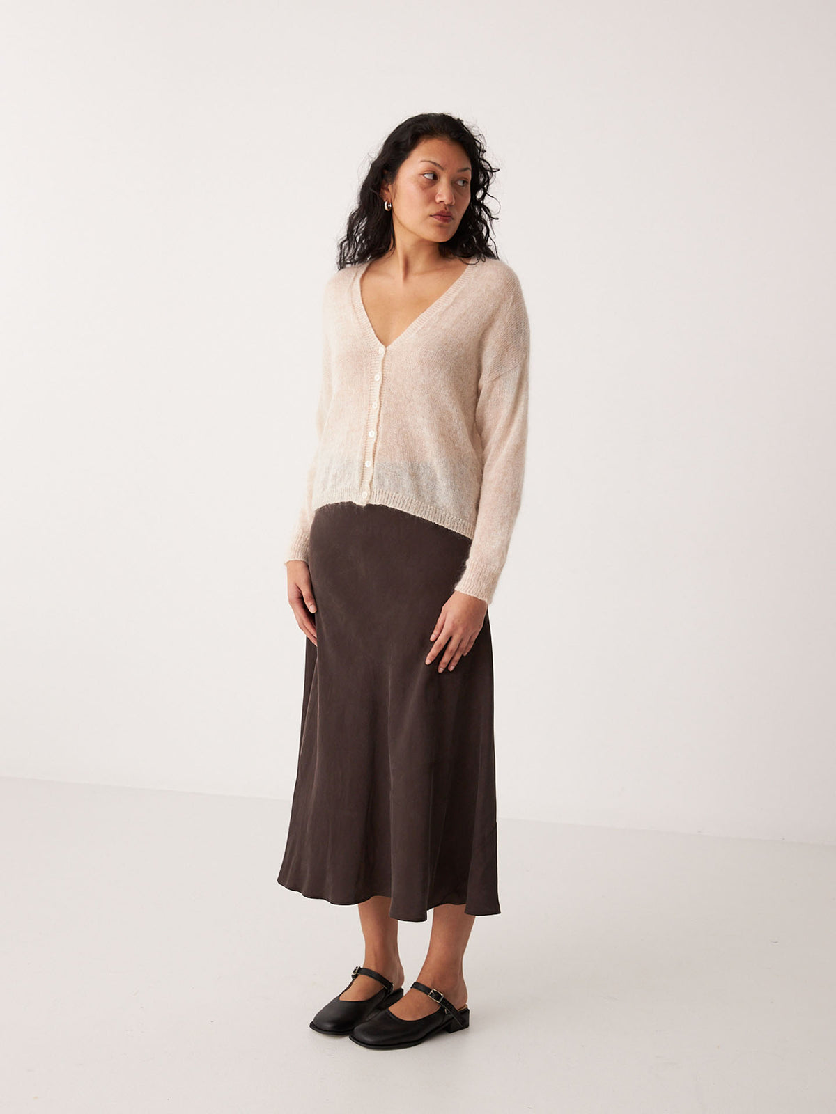 A petite woman stands against a plain white background wearing the Mist Cardigan – Creme, a dark brown midi skirt, and black Mary Jane shoes.