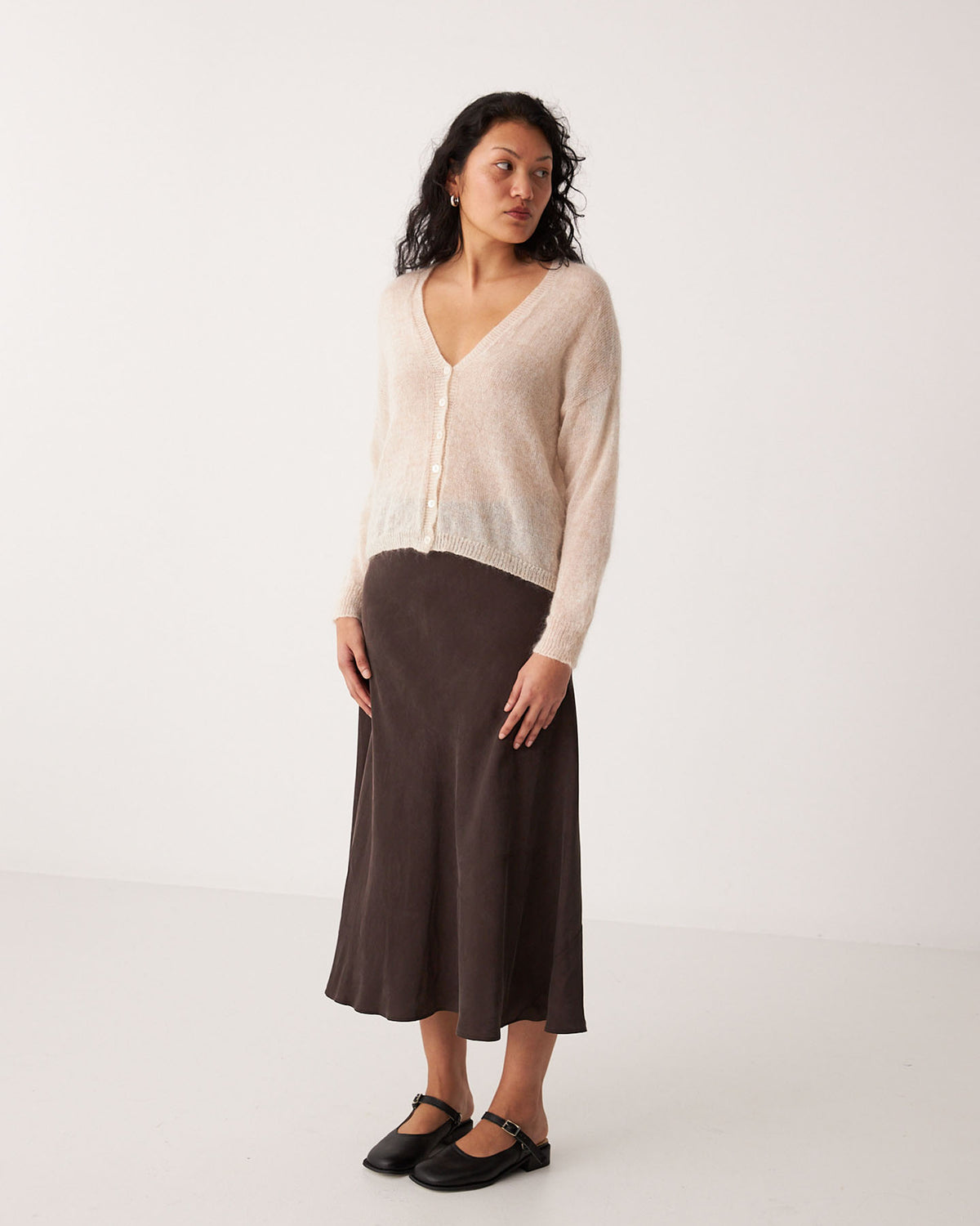 A petite woman stands against a plain white background wearing the Mist Cardigan – Creme, a dark brown midi skirt, and black Mary Jane shoes.