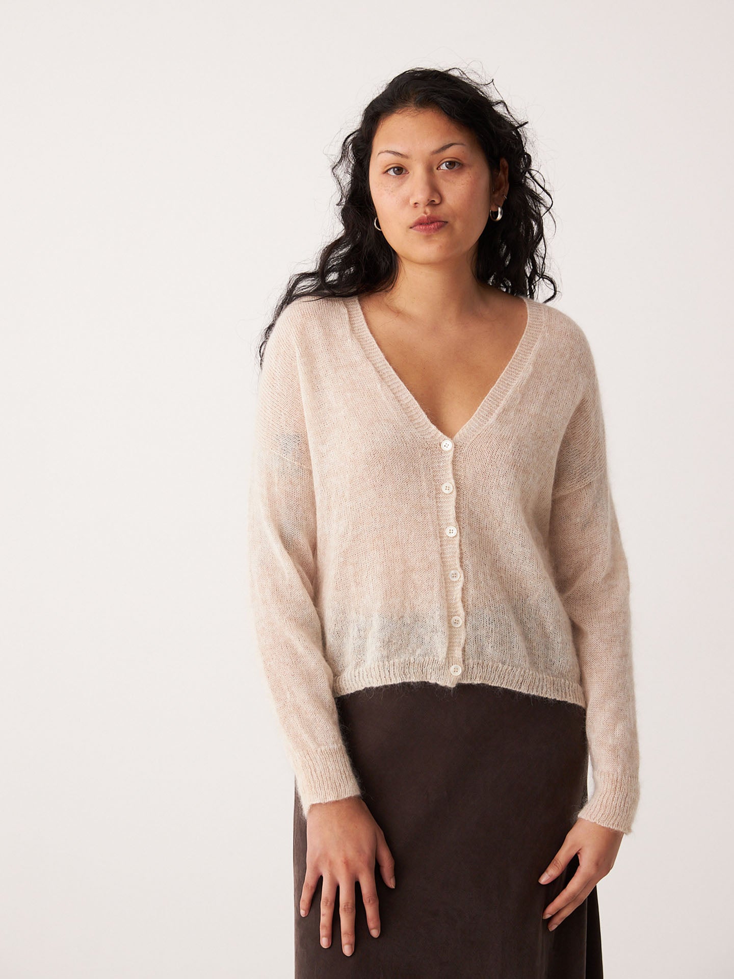 A relaxed woman with long, wavy dark hair wears the Mist Cardigan in Creme and a dark skirt, standing against a plain light background.
