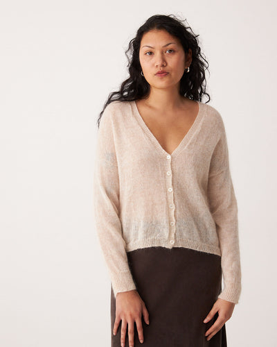 A relaxed woman with long, wavy dark hair wears the Mist Cardigan in Creme and a dark skirt, standing against a plain light background.