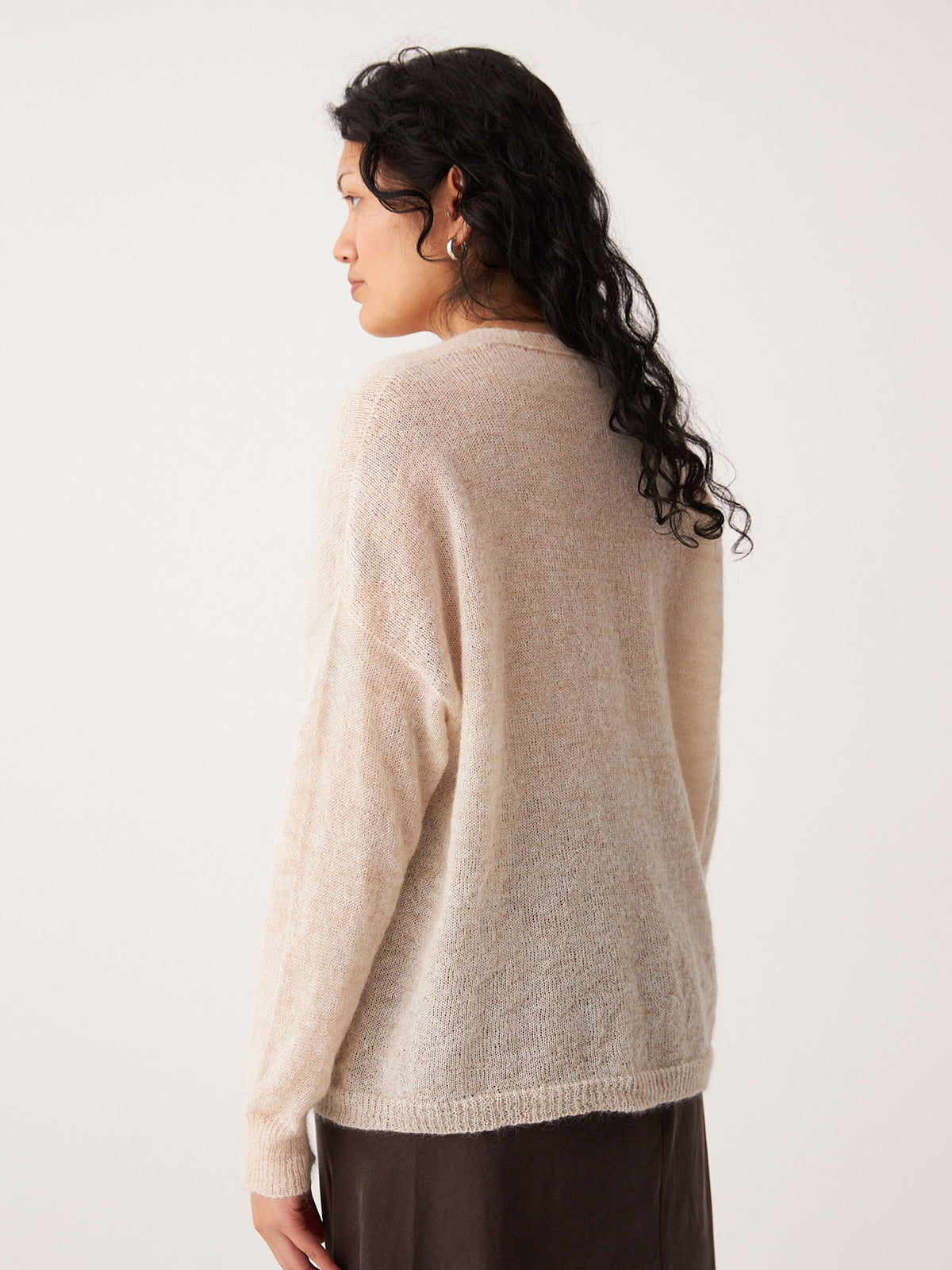 A person with long, wavy dark hair stands facing away, wearing the Mist Cardigan – Creme in petite size with a dark skirt against a plain background.