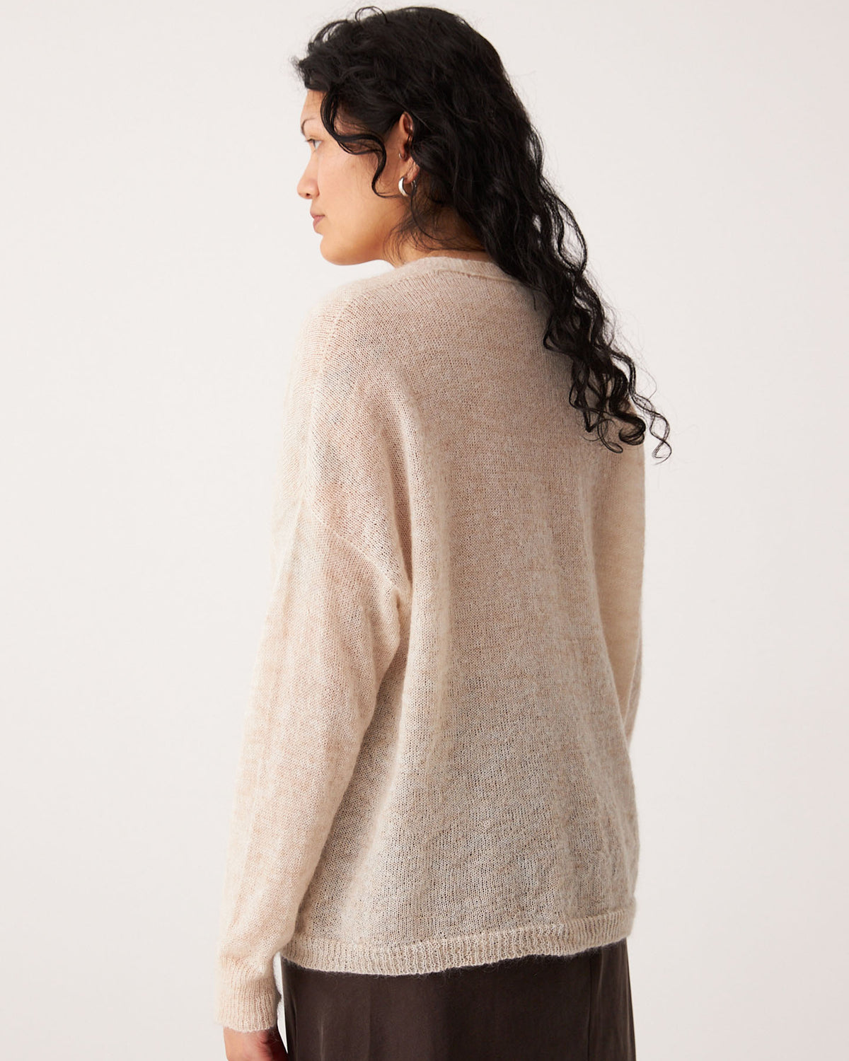 A person with long, wavy dark hair stands facing away, wearing the Mist Cardigan – Creme in petite size with a dark skirt against a plain background.