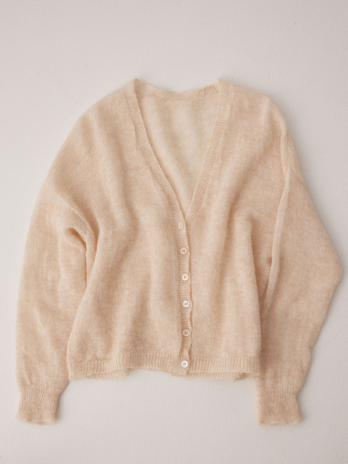 The Mist Cardigan – Creme is a beige, cropped, long-sleeved cardigan with a relaxed fit, V-neck, and white buttons, shown laid flat on a light surface.