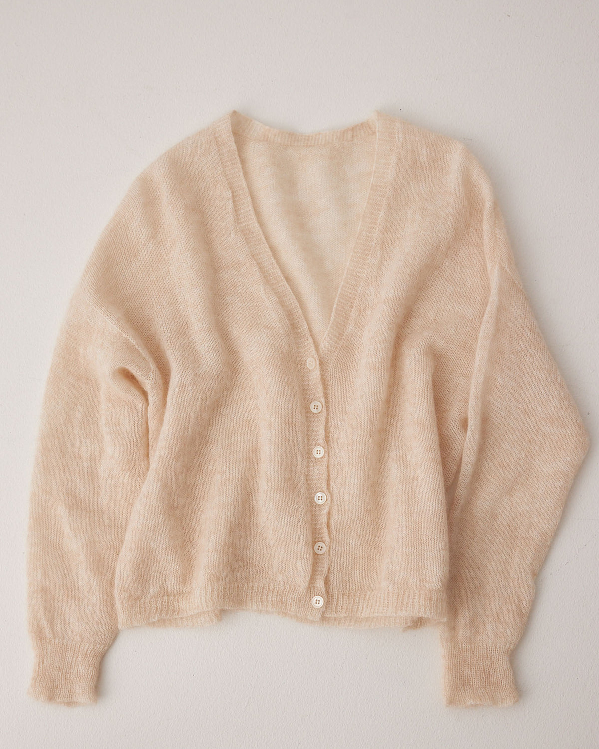 The Mist Cardigan – Creme is a beige, cropped, long-sleeved cardigan with a relaxed fit, V-neck, and white buttons, shown laid flat on a light surface.