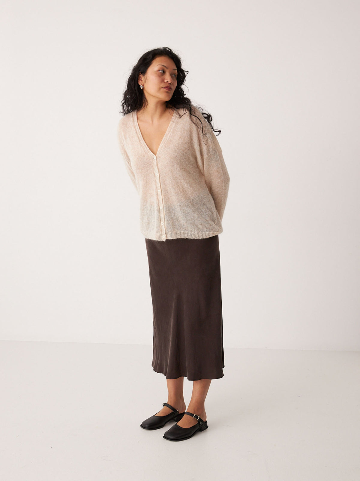 A petite woman stands in a neutral space wearing the Mist Cardigan – Creme, a dark brown midi skirt, and black Mary Jane shoes.