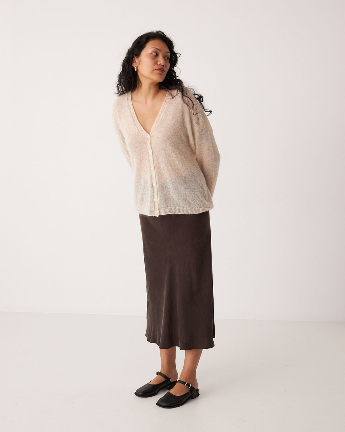 A petite woman stands in a neutral space wearing the Mist Cardigan – Creme, a dark brown midi skirt, and black Mary Jane shoes.