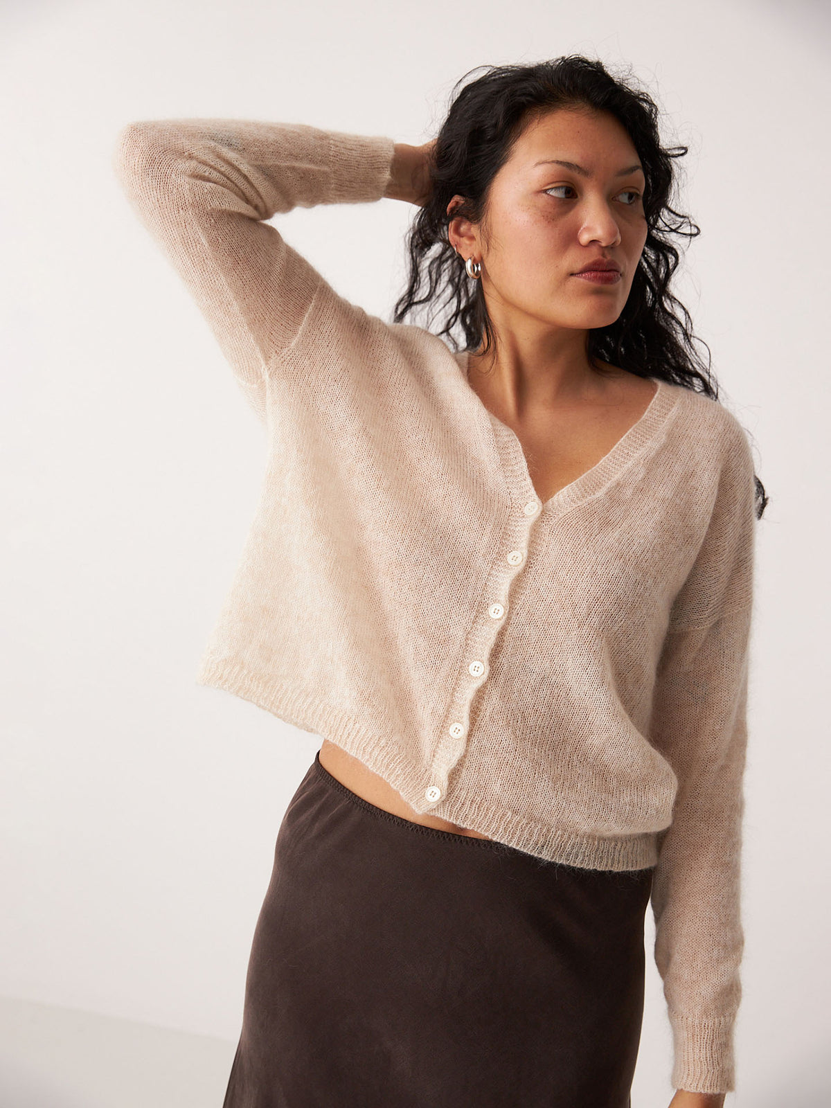 Petite woman wearing the Mist Cardigan – Creme, a relaxed beige button-up, paired with a dark skirt and posing against a plain light background with one arm raised behind her head.