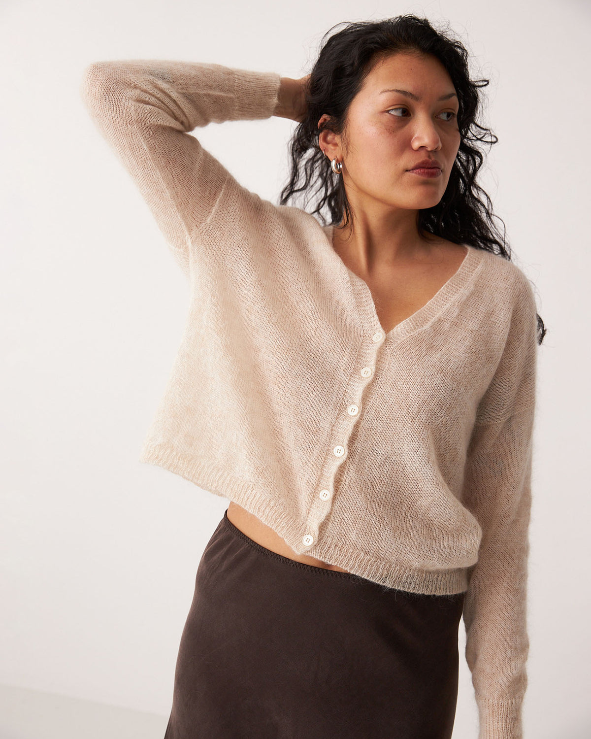 Petite woman wearing the Mist Cardigan – Creme, a relaxed beige button-up, paired with a dark skirt and posing against a plain light background with one arm raised behind her head.