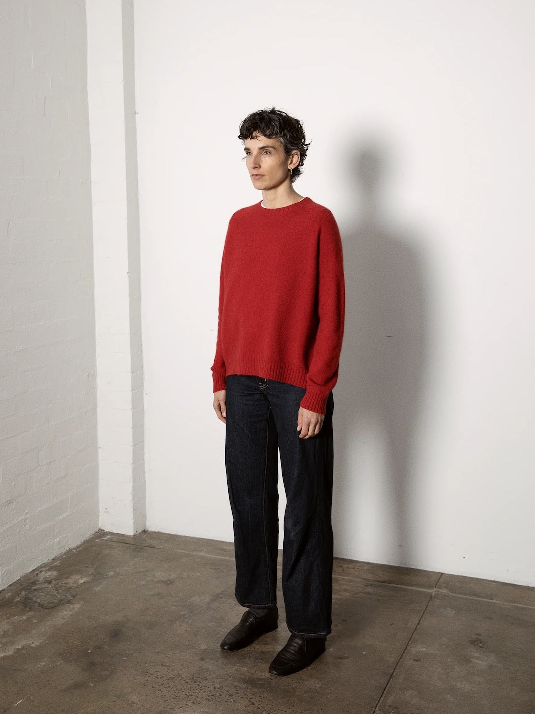 A petite person with short hair stands against a plain white wall on a concrete floor, wearing the fitted Nimbus Raglan Knit – Poppy by Francie and dark pants.