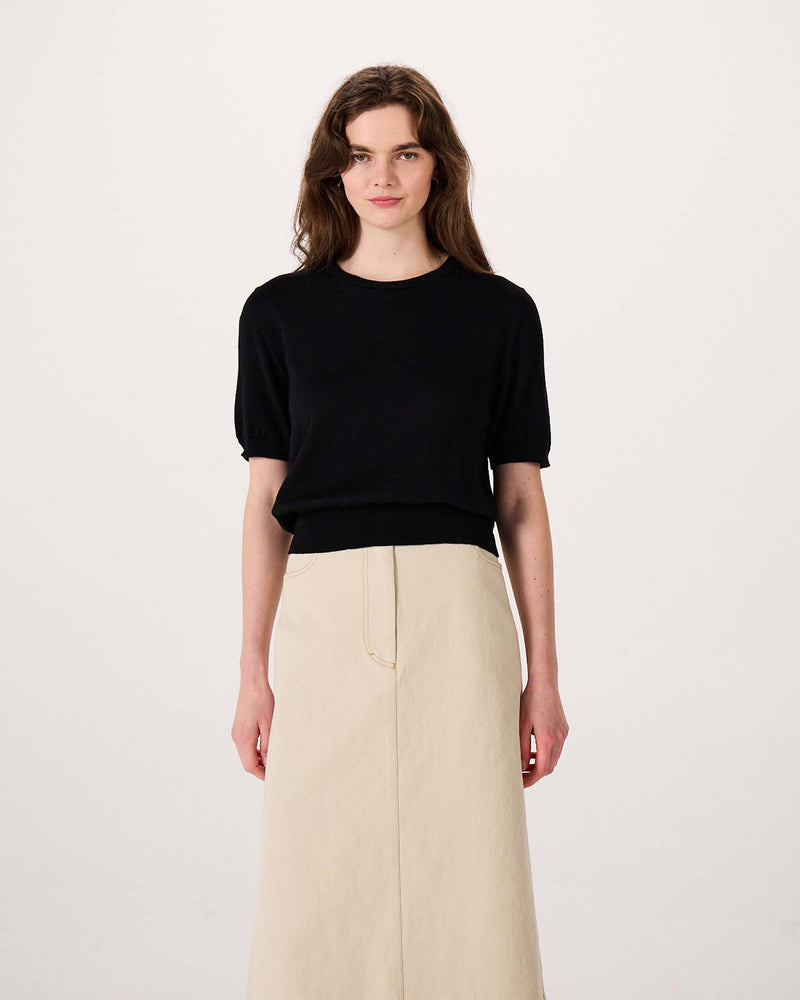Woman standing against a plain white background, wearing a black short-sleeve top and a high-waisted beige skirt.