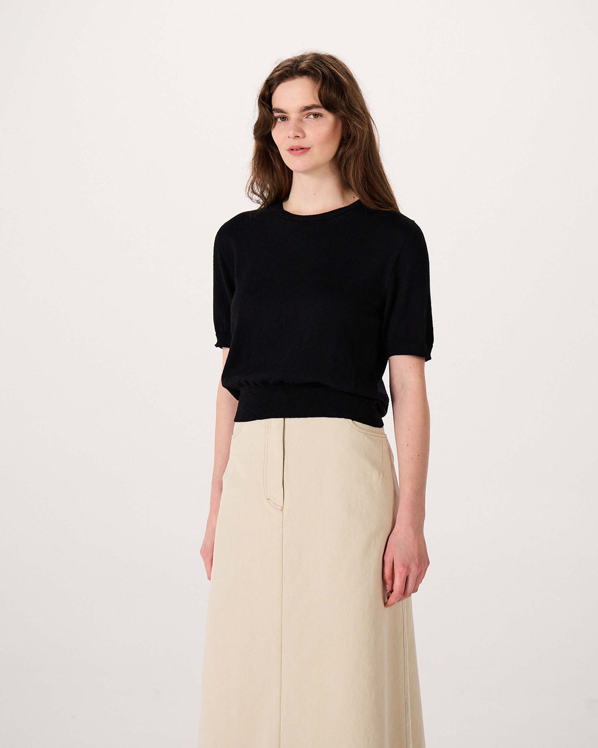 A person stands against a plain background wearing a black short-sleeve top and a long beige skirt.