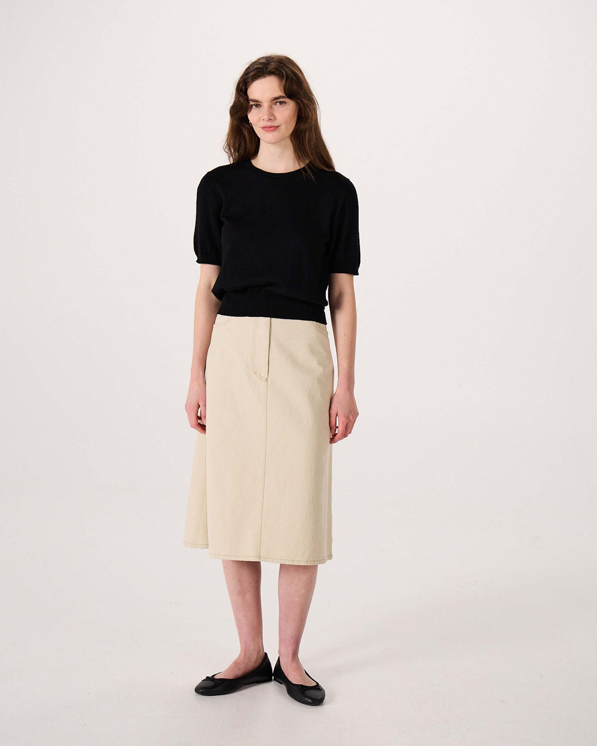 Woman standing against a plain background, wearing a black short-sleeve top, beige knee-length skirt, and black flats.