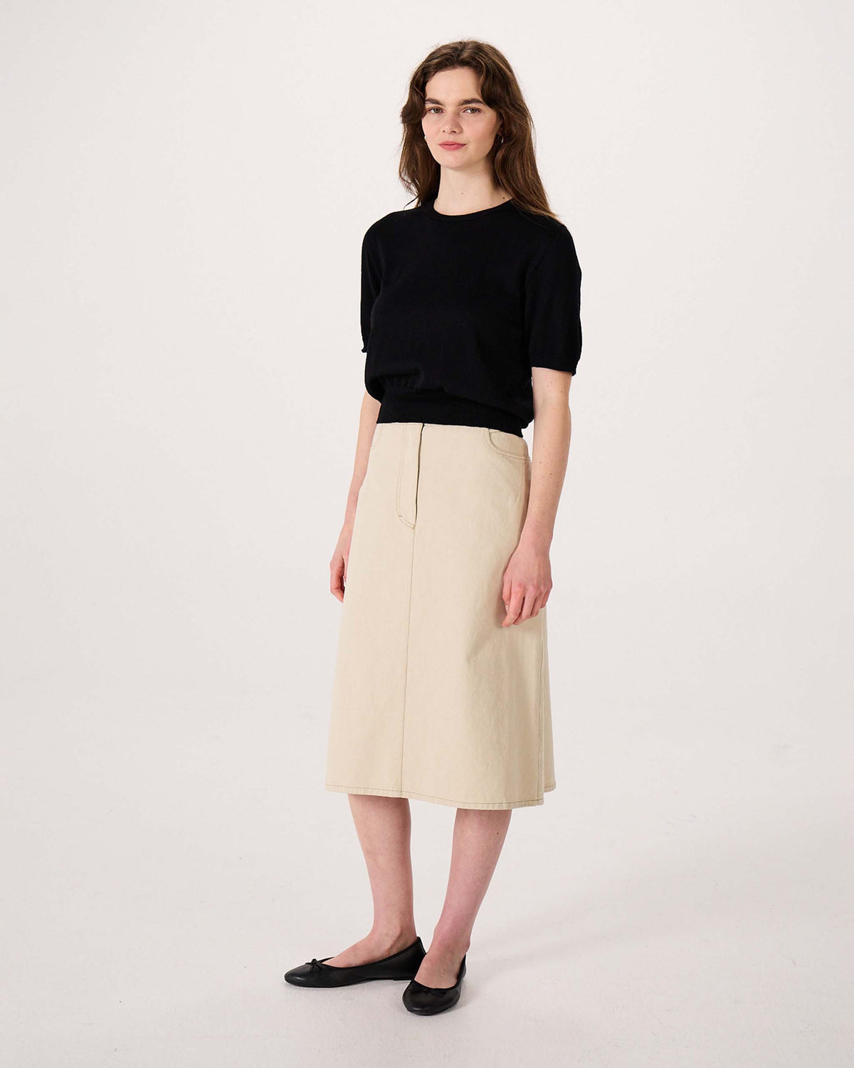 A woman stands facing forward wearing a black short-sleeve top, beige knee-length skirt, and black flats against a plain white background.