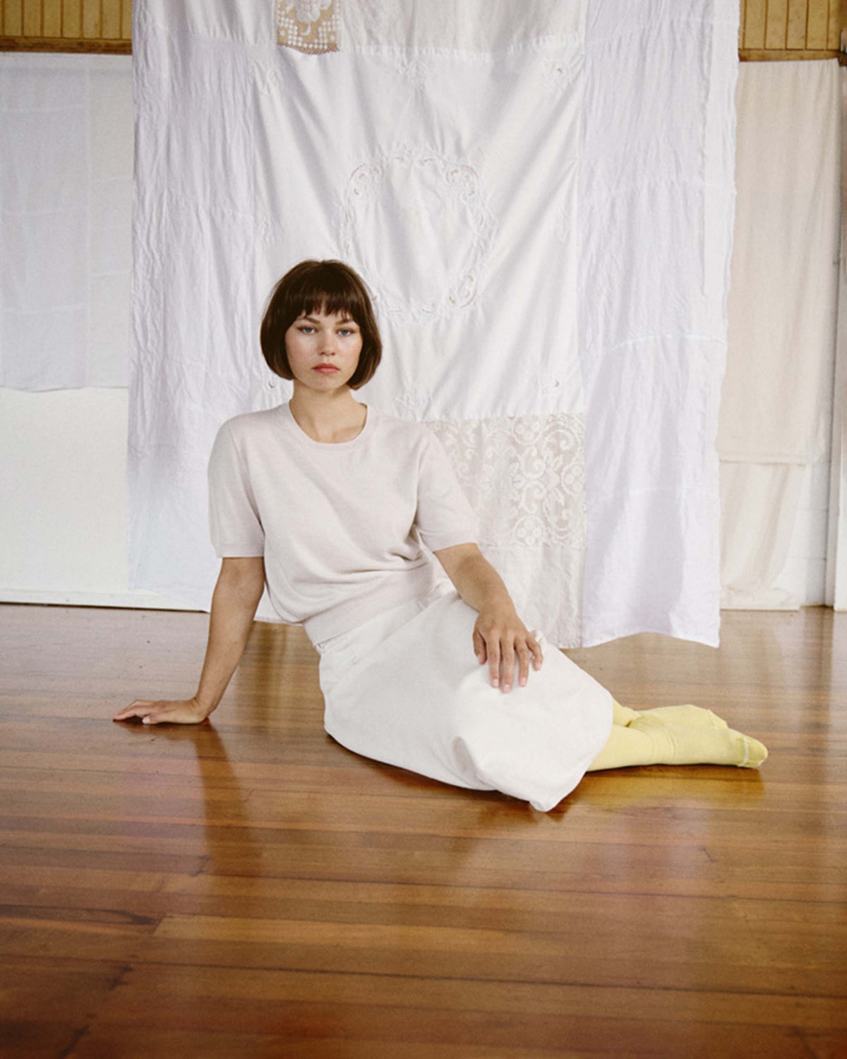 A person in a white outfit and yellow socks sits on a wooden floor in front of hanging white fabric.