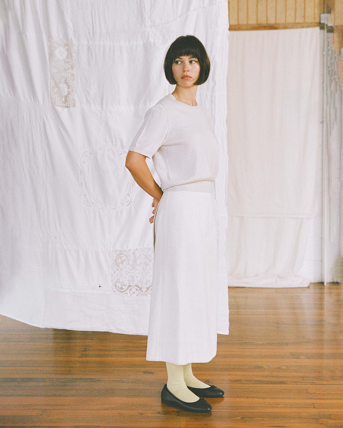 Woman with short dark hair stands indoors, wearing a white top, white skirt, yellow socks, and black shoes. She poses with hands behind her back against a white fabric backdrop.