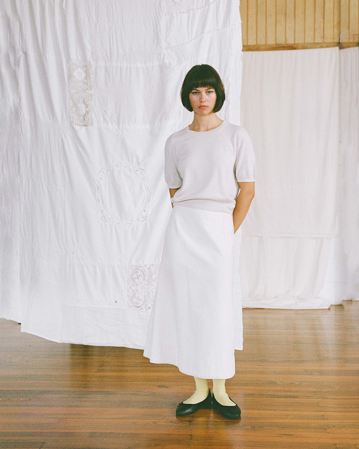 A woman stands indoors on a wooden floor, wearing a light short-sleeve top, a long white skirt, yellow socks, and black shoes, with white fabric draped in the background.