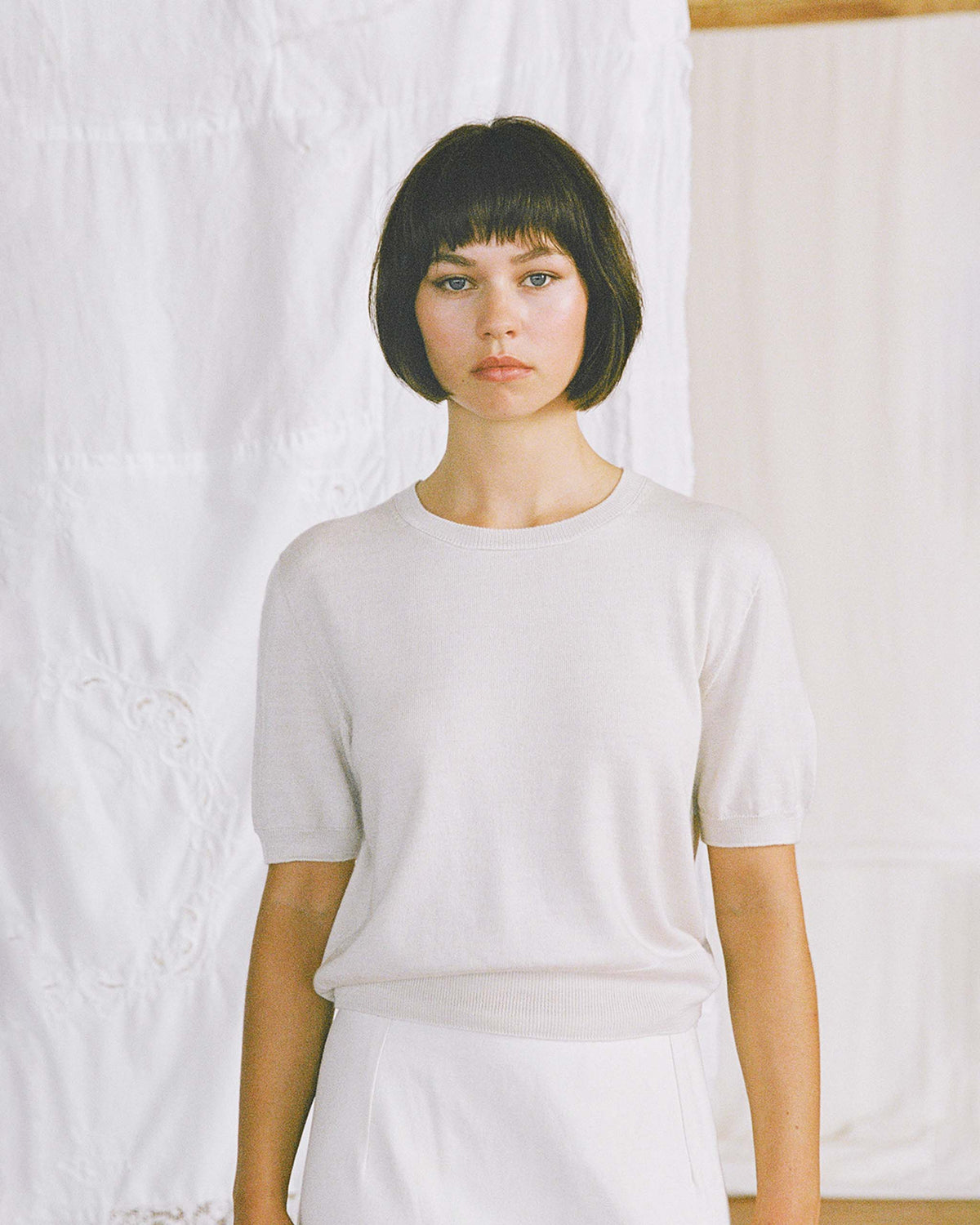 Woman with short dark hair wearing a light-colored short-sleeve sweater and skirt stands in front of a white textured background.
