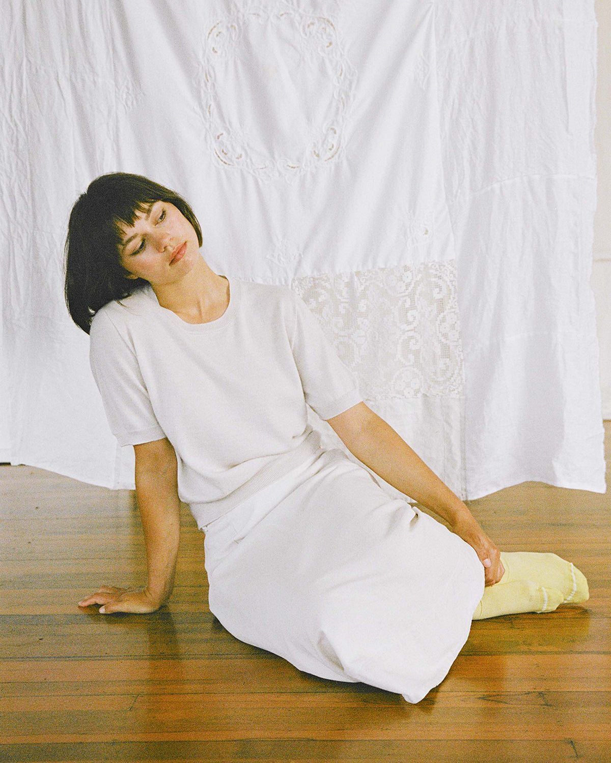 A woman in a white outfit sits on a wooden floor with her head tilted and eyes closed, in front of a white fabric backdrop.