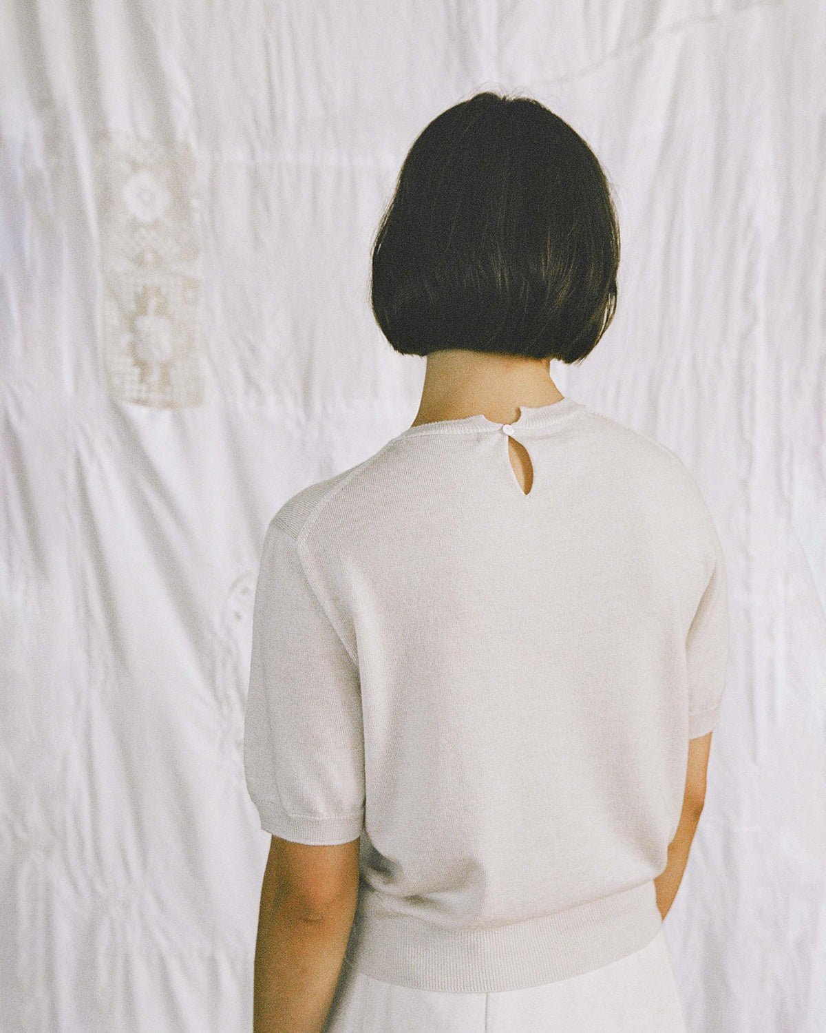 Person with short dark hair facing away, wearing a light-colored short-sleeve top, standing in front of a white textured background.