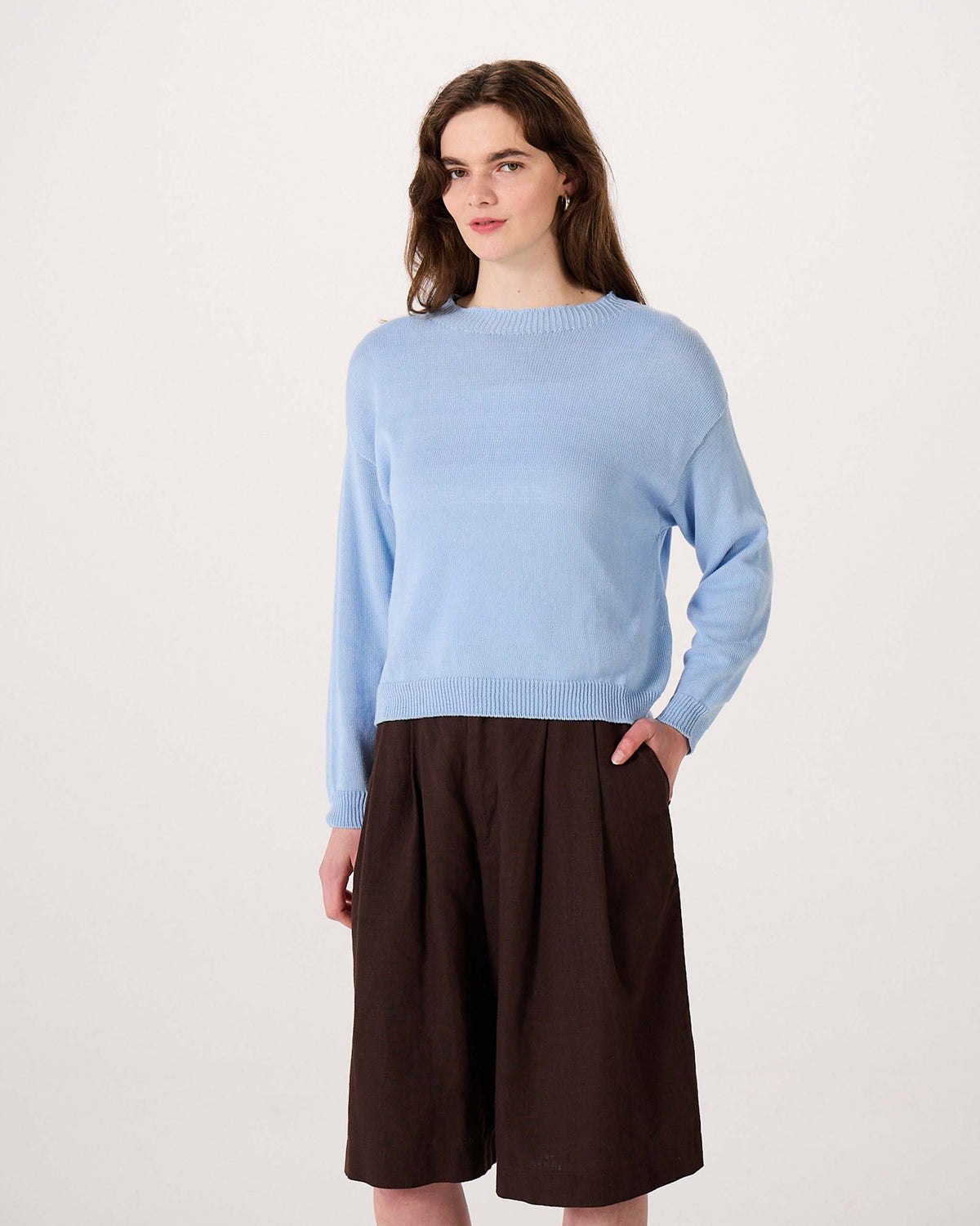 Woman wearing a light blue sweater and brown knee-length wide-leg shorts stands against a plain white background with one hand in her pocket.