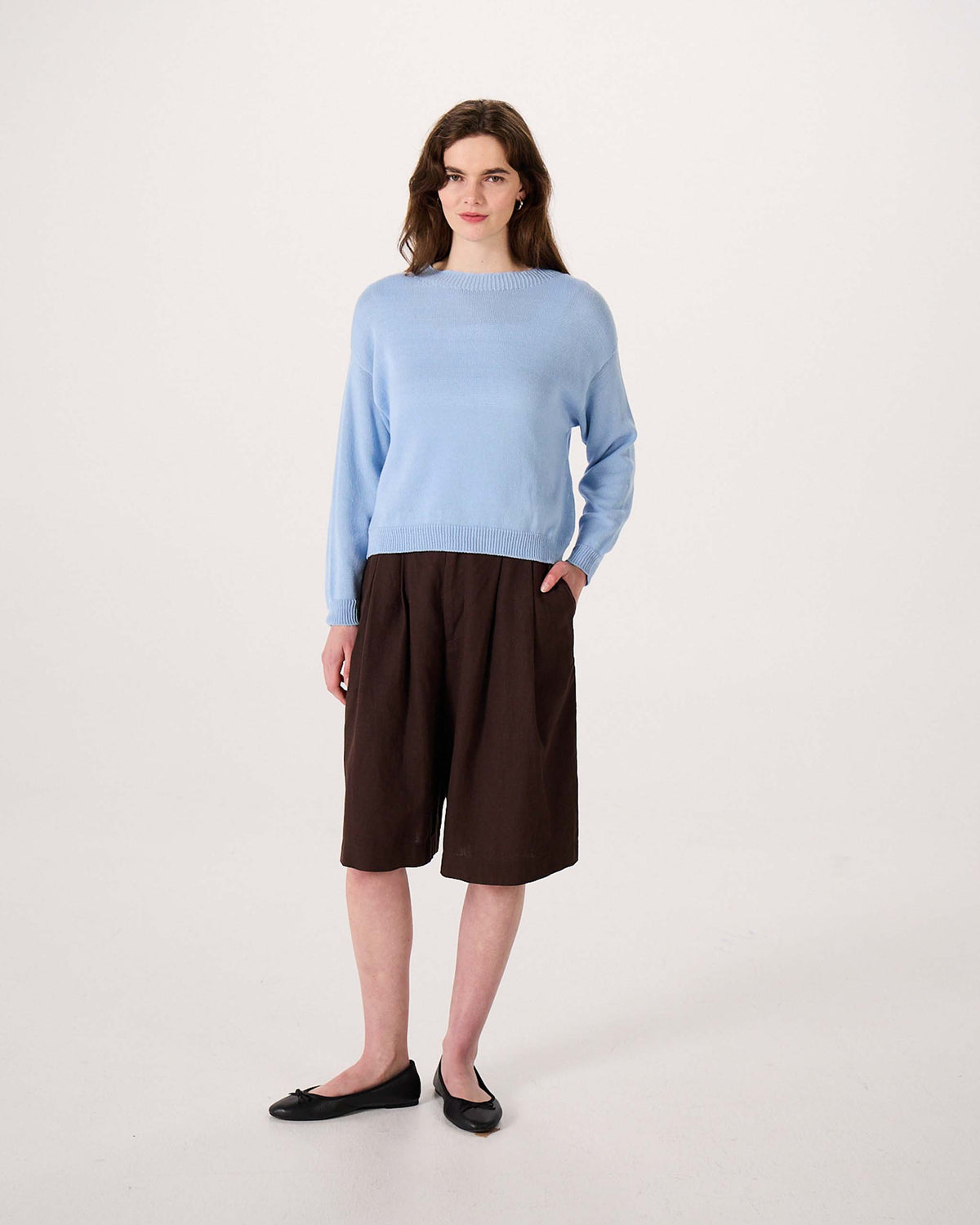 A woman stands wearing a light blue sweater, dark brown knee-length shorts, and black flats against a plain white background.