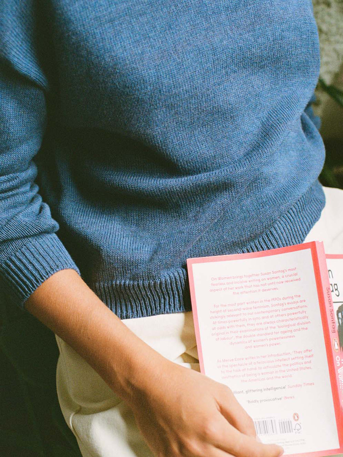 A person in a relaxed fit Pebble Knit – Fiord Blue sweater and light pants is holding a red-covered book, displaying its back.