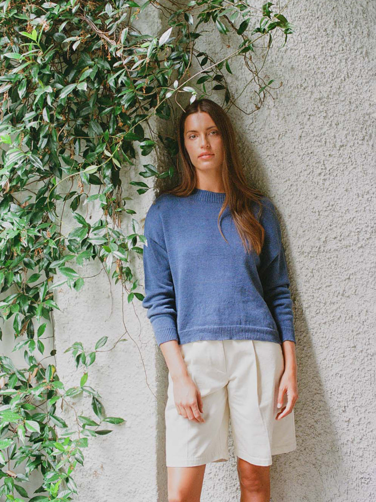 A petite woman with long brown hair stands against a textured white wall beside leafy vines, wearing the Pebble Knit – Fiord Blue cropped sweater and white shorts.