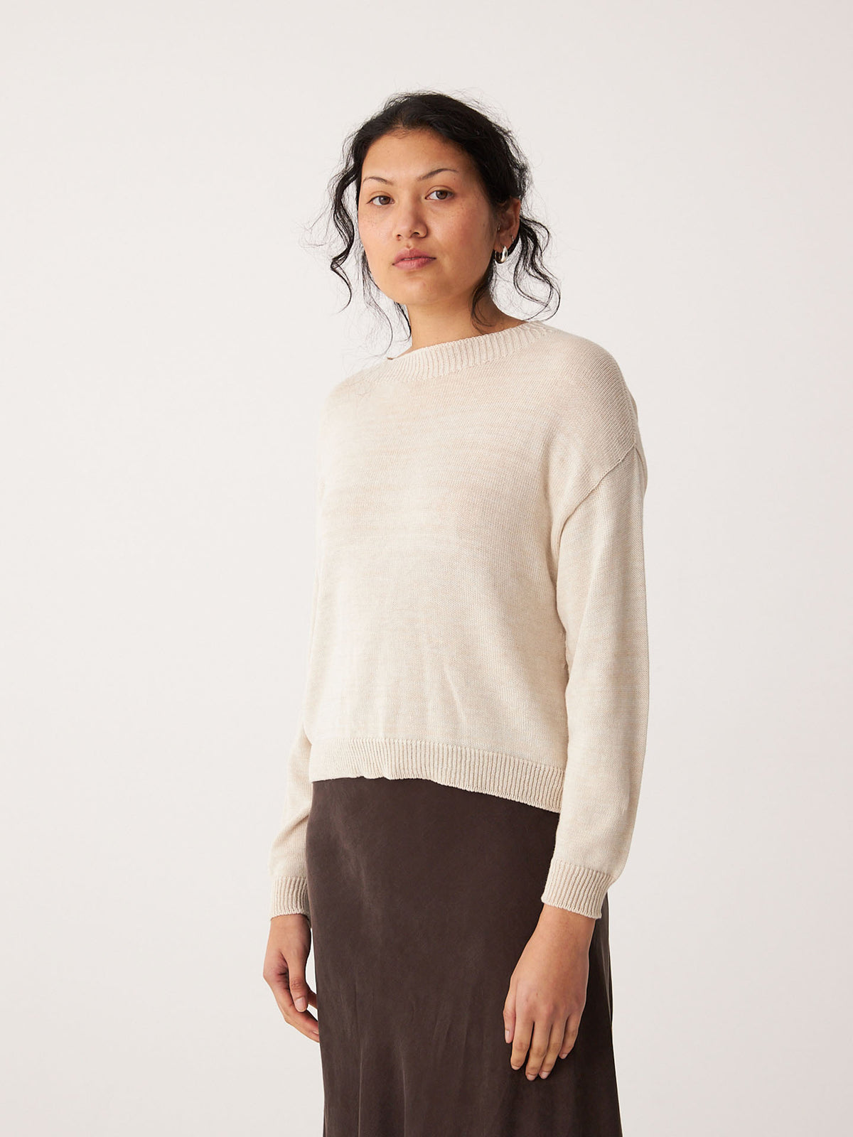 A person wears the Pebble Knit – Creme, a light beige relaxed fit long-sleeve sweater, paired with a dark brown skirt, standing against a plain white background.