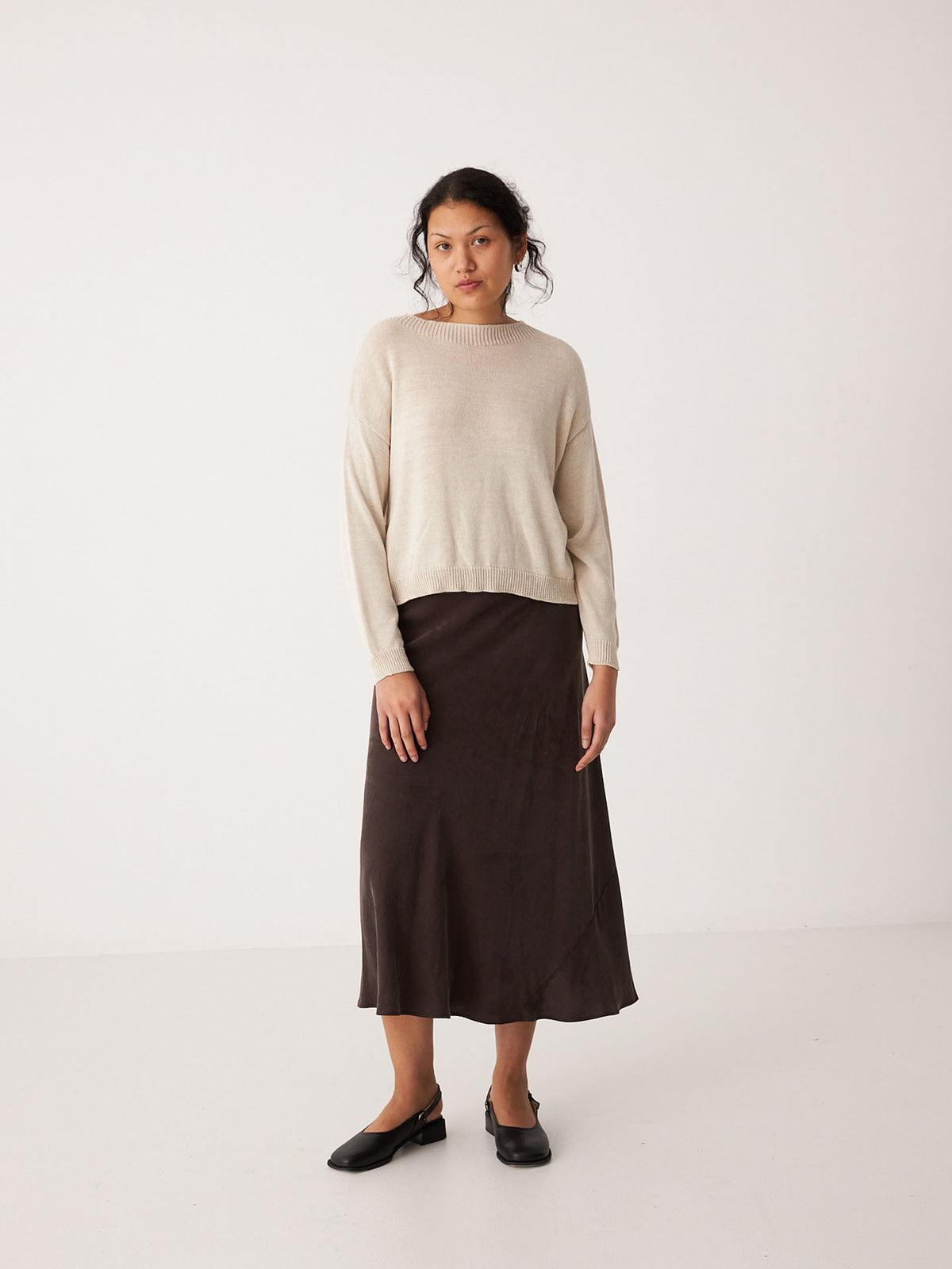 A woman stands against a plain white background, wearing the Pebble Knit – Creme relaxed fit sweater, paired with a dark brown midi skirt and black shoes.