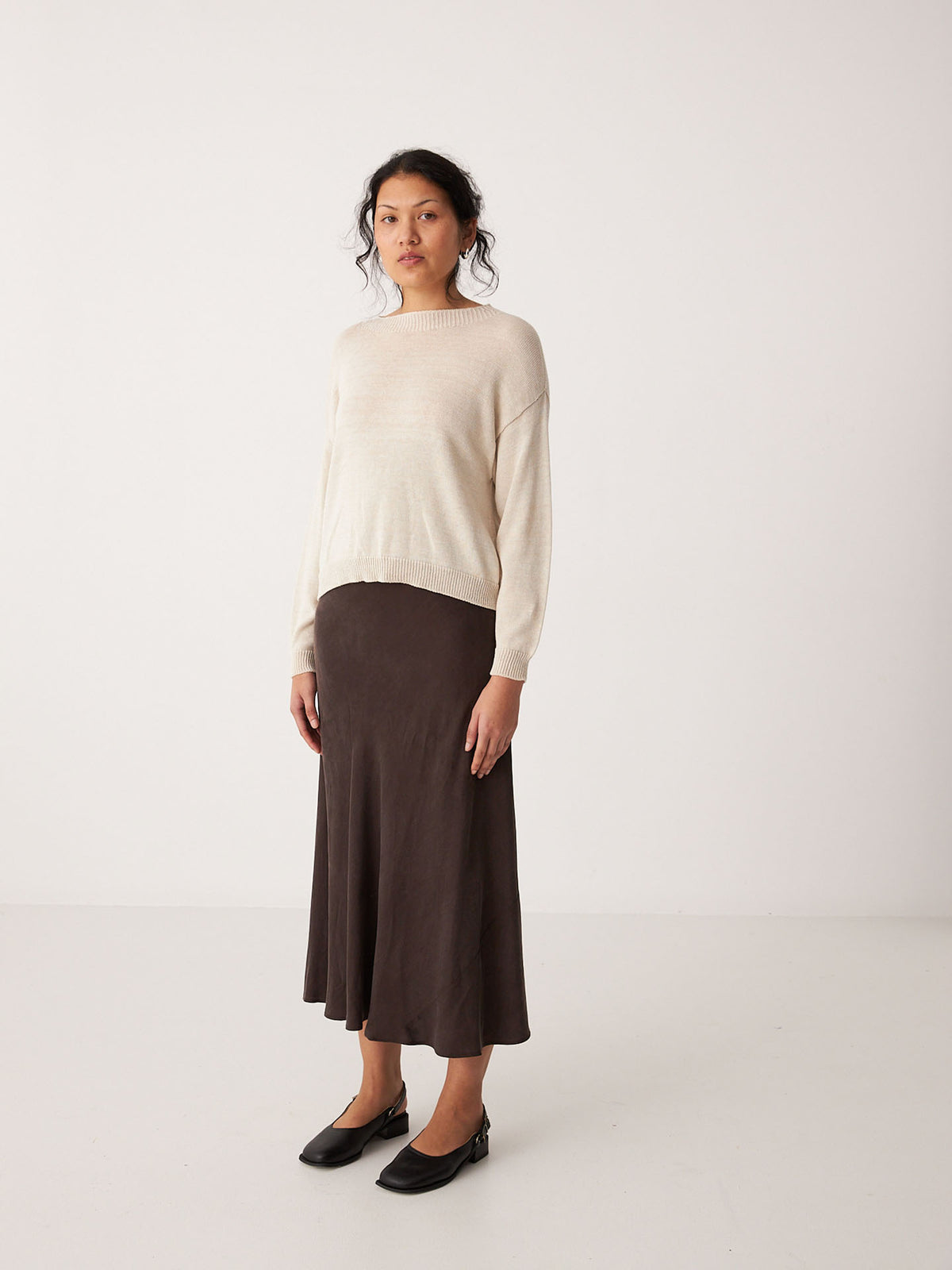 A person stands in a plain room wearing the Pebble Knit – Creme sweater, a dark brown midi skirt, and black flat shoes, looking towards the camera.