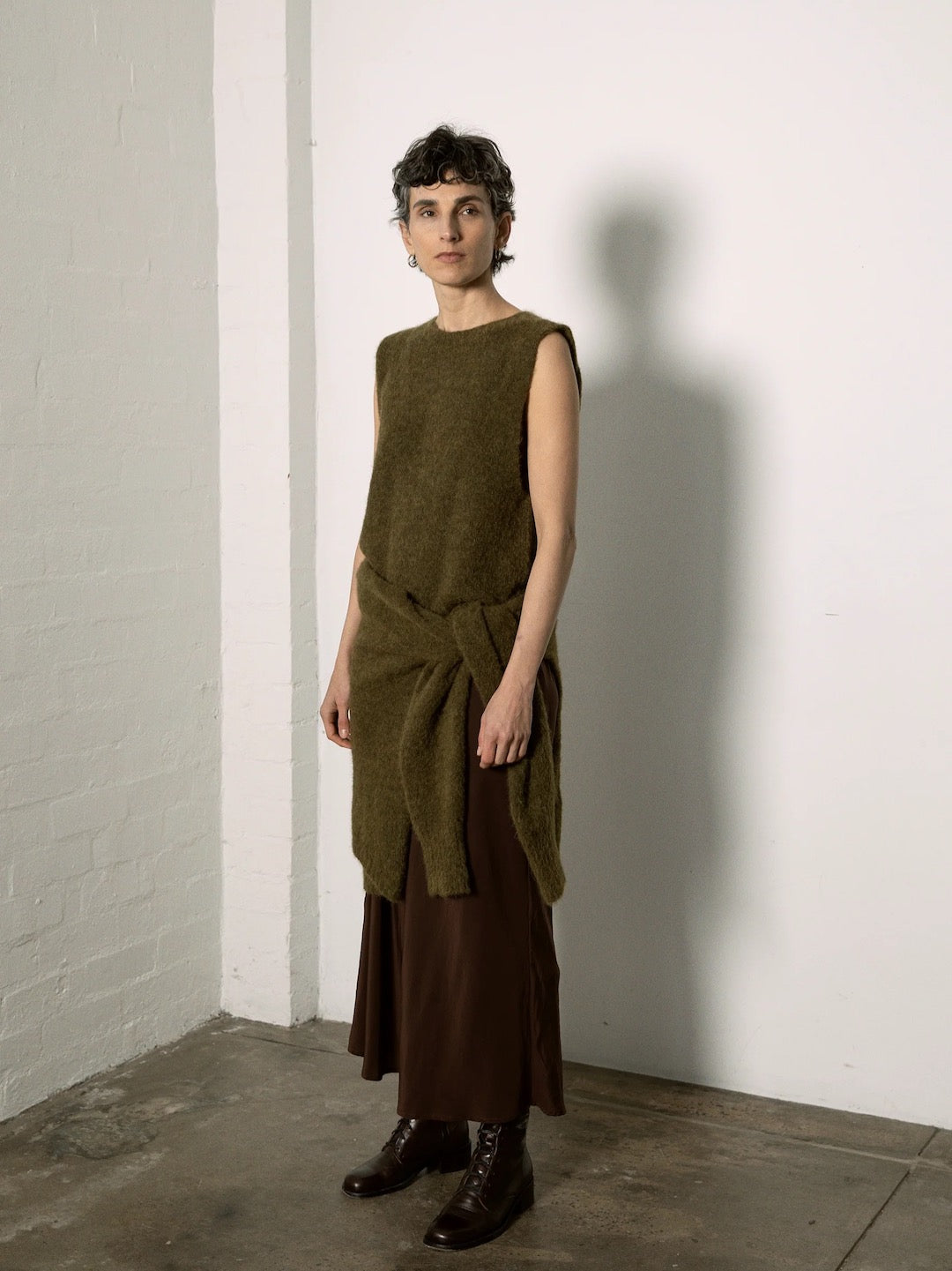 A petite person stands by a white wall, showcasing the fitted style of Francie's Picnic Vest in Moss. Their short hair adds an edgy touch, paired perfectly with a long skirt and dark boots.