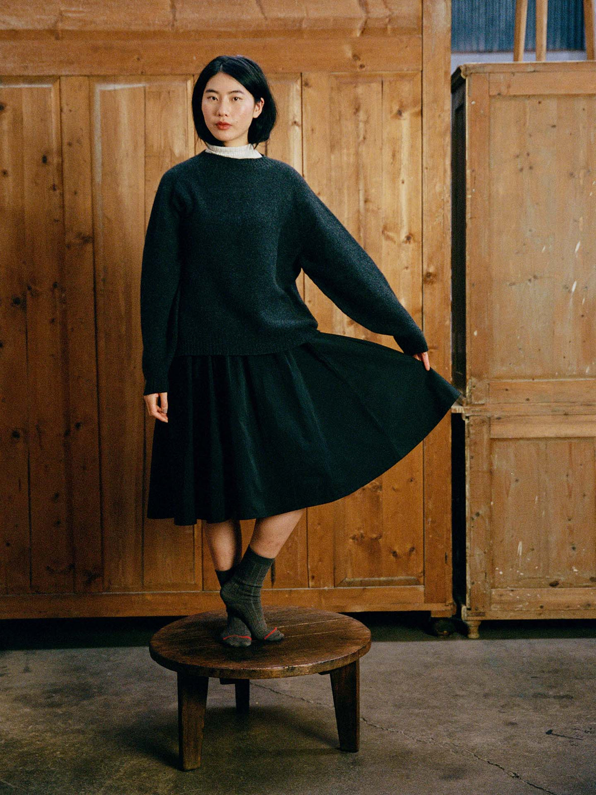 A woman wears the Poet Knit – Almost Black, styled with a black skirt and gray socks, standing on a round wooden stool in front of wooden cabinets.