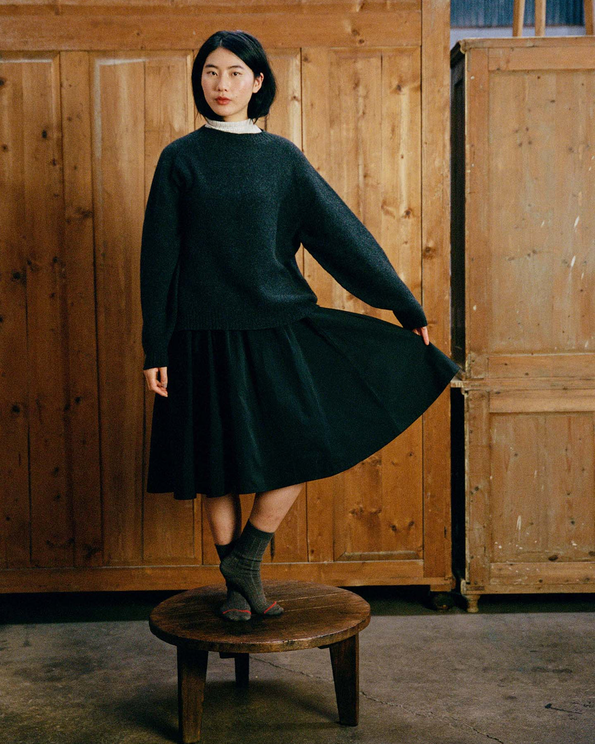 A woman wears the Poet Knit – Almost Black, styled with a black skirt and gray socks, standing on a round wooden stool in front of wooden cabinets.