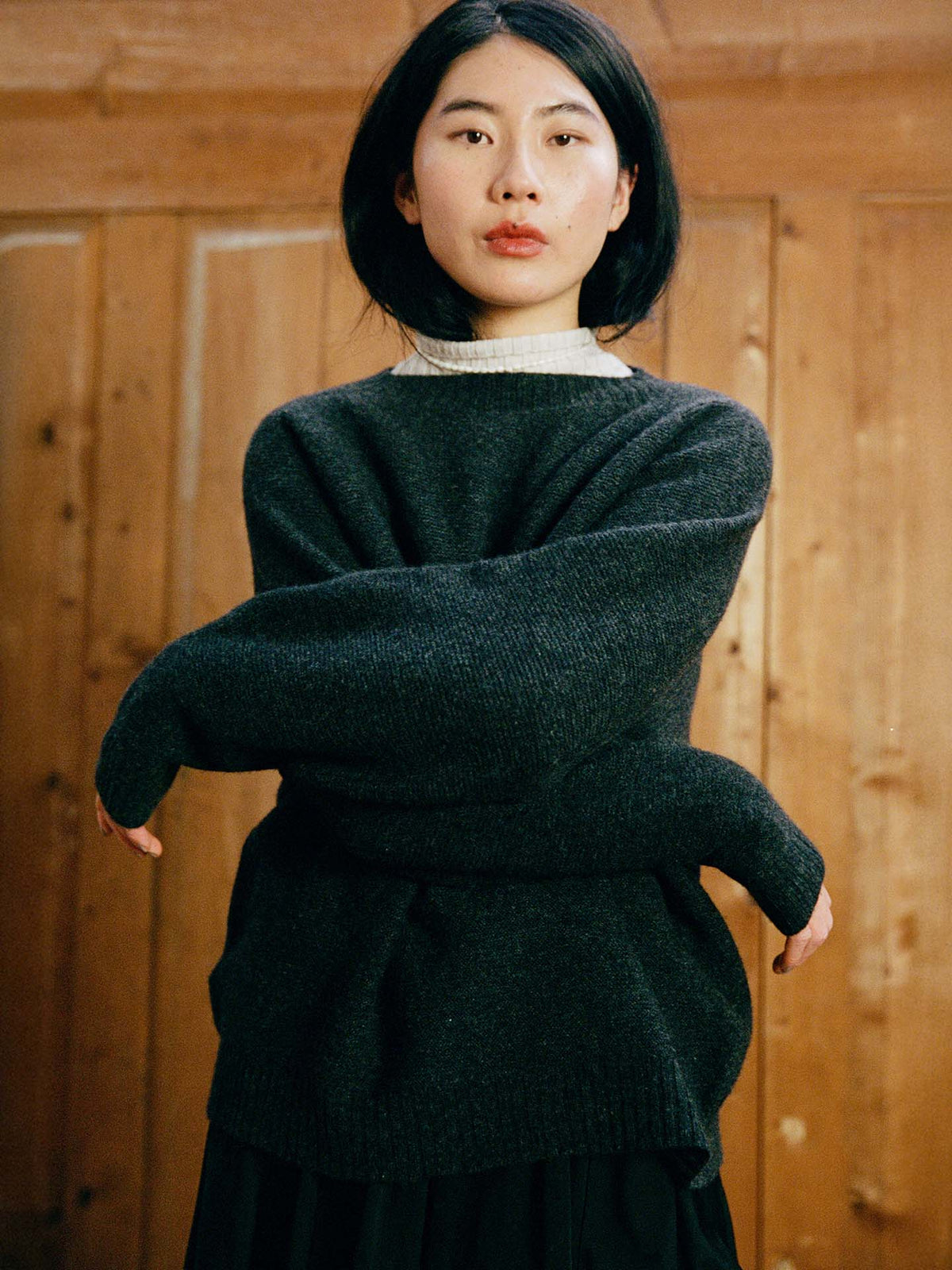 A woman with straight black hair and a neutral expression stands indoors against wooden paneling, wearing the relaxed fit Poet Knit – Almost Black sweater over a light turtleneck.