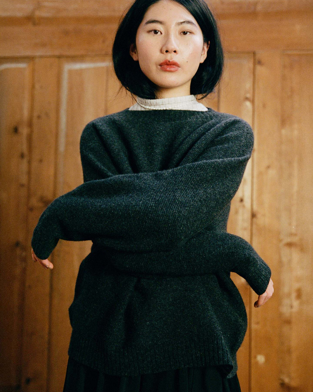 A woman with straight black hair and a neutral expression stands indoors against wooden paneling, wearing the relaxed fit Poet Knit – Almost Black sweater over a light turtleneck.