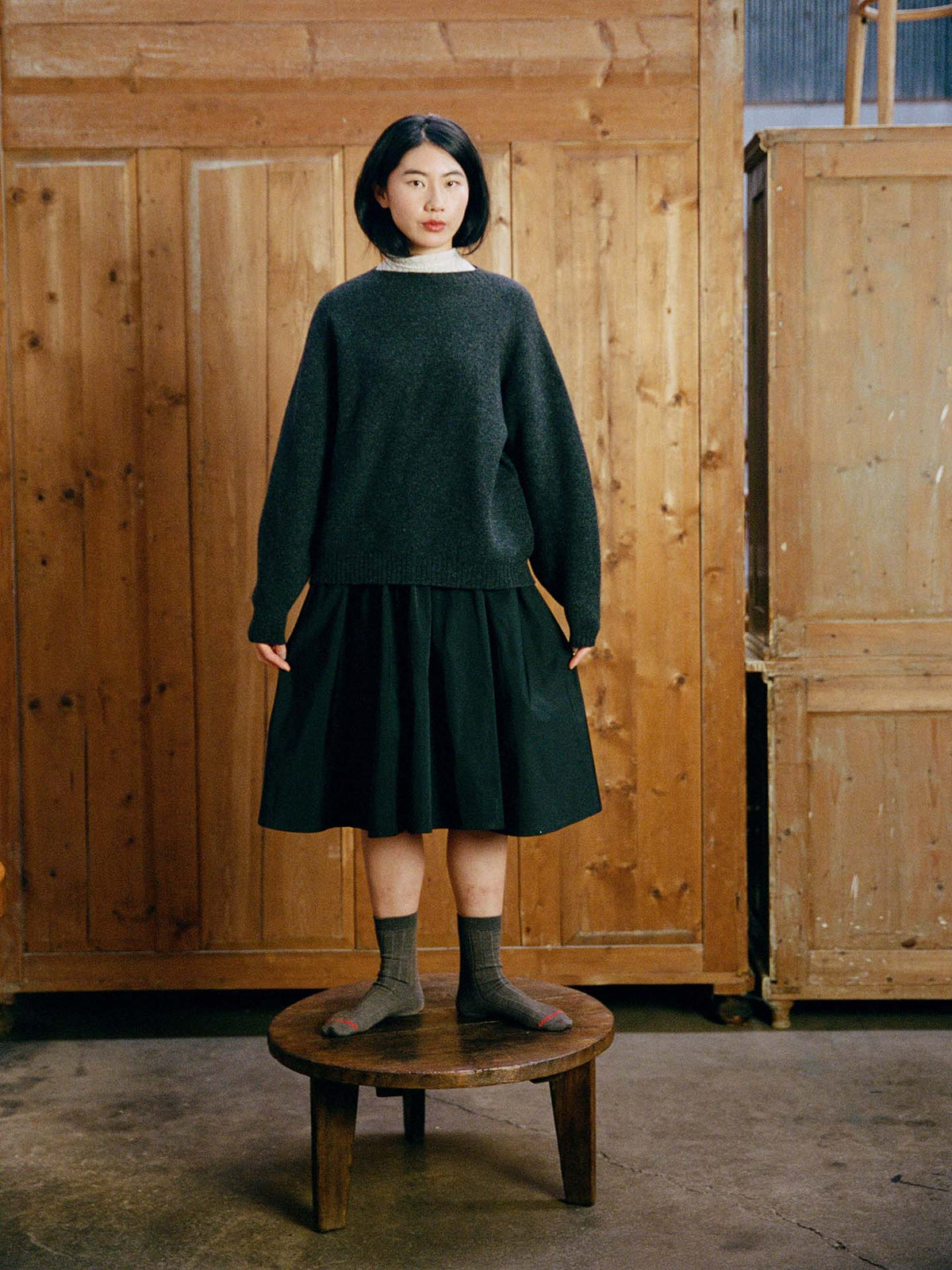 A woman in the relaxed fit Poet Knit – Almost Black sweater, black skirt, and gray socks stands on a round wooden stool in front of wooden paneling.