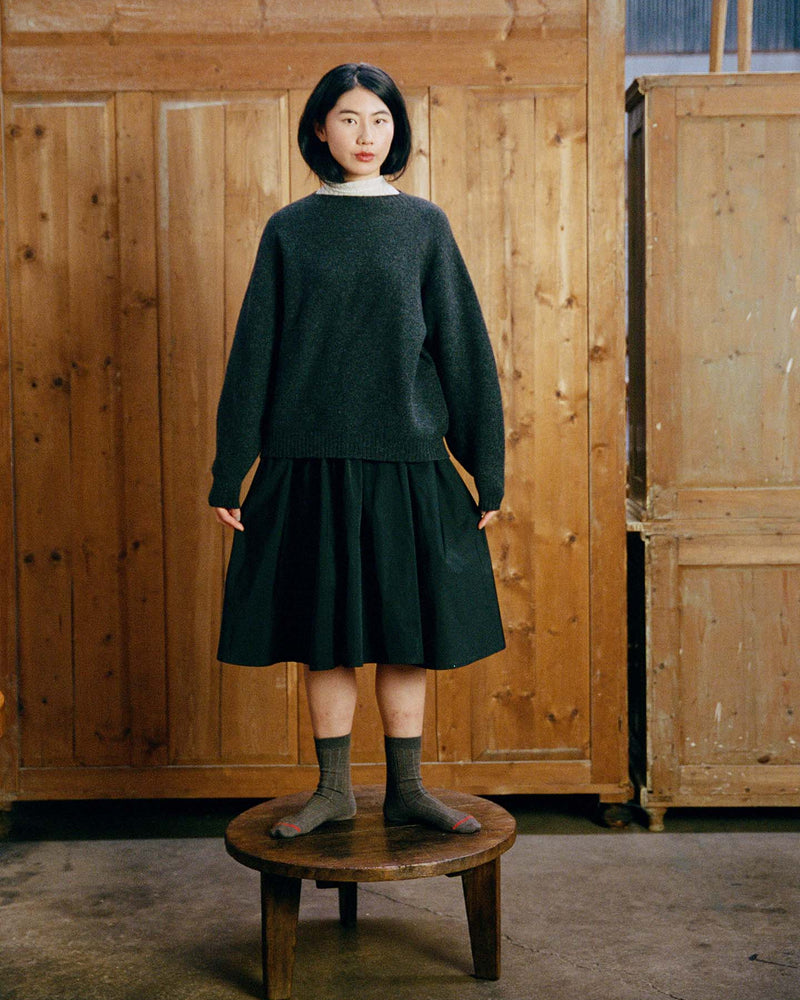 A woman in the relaxed fit Poet Knit – Almost Black sweater, black skirt, and gray socks stands on a round wooden stool in front of wooden paneling.