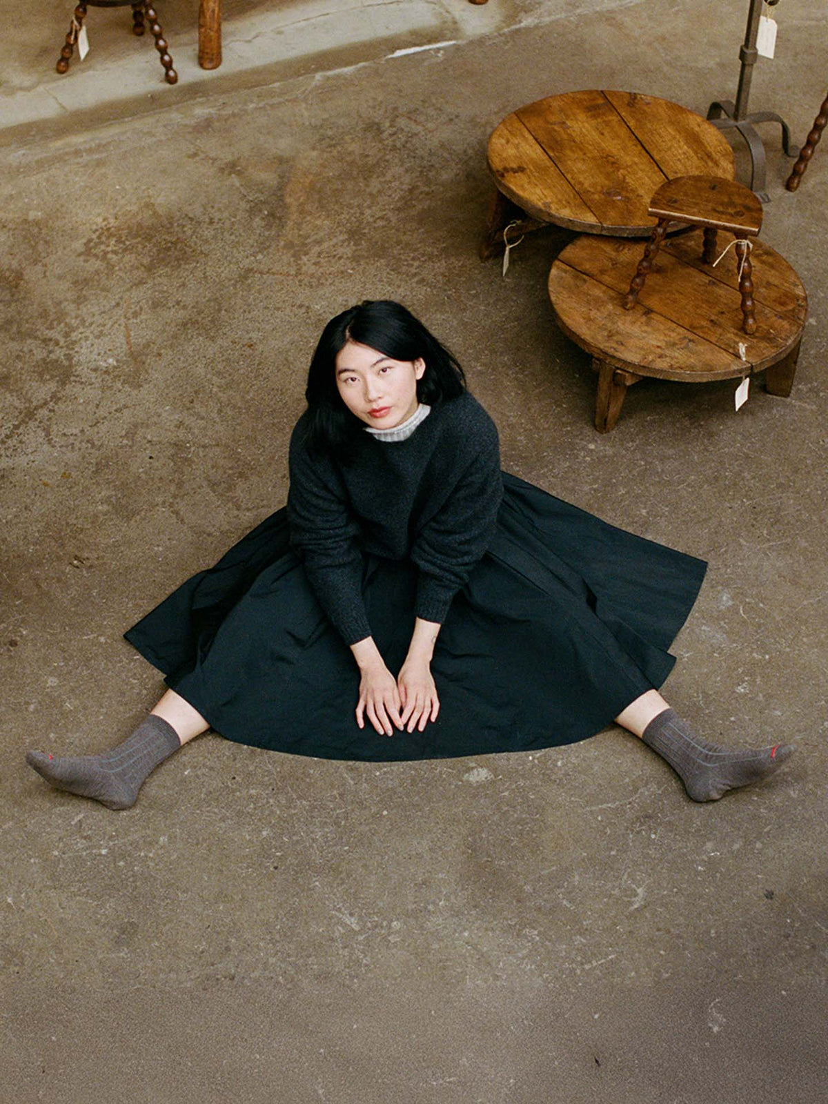 A woman with dark hair sits on the floor with legs in a V shape, wearing the Poet Knit in Almost Black and gray socks, viewed from above. Wooden stools are nearby.