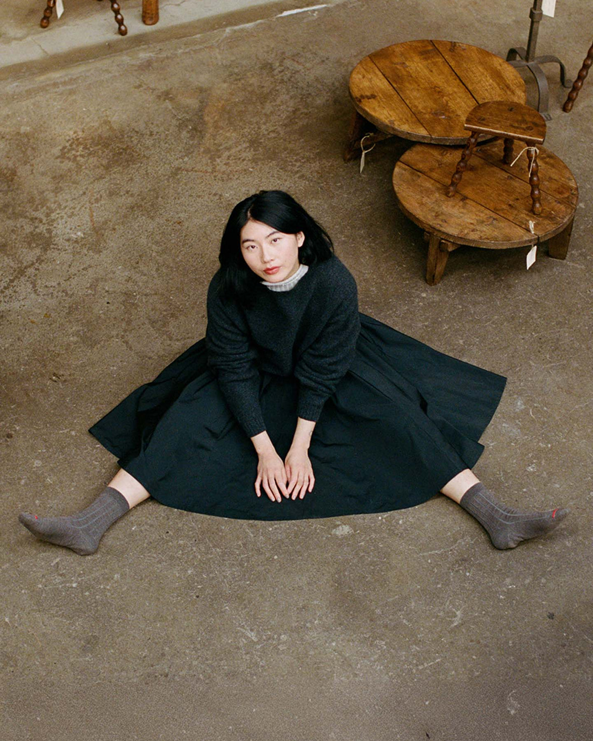 A woman with dark hair sits on the floor with legs in a V shape, wearing the Poet Knit in Almost Black and gray socks, viewed from above. Wooden stools are nearby.