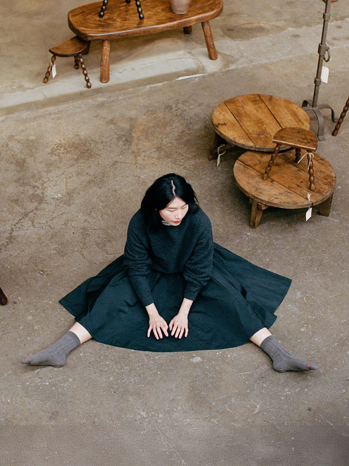 A woman with black hair sits on a concrete floor wearing the Poet Knit – Almost Black and gray socks, her legs stretched out and hands on her knees, surrounded by small wooden tables.