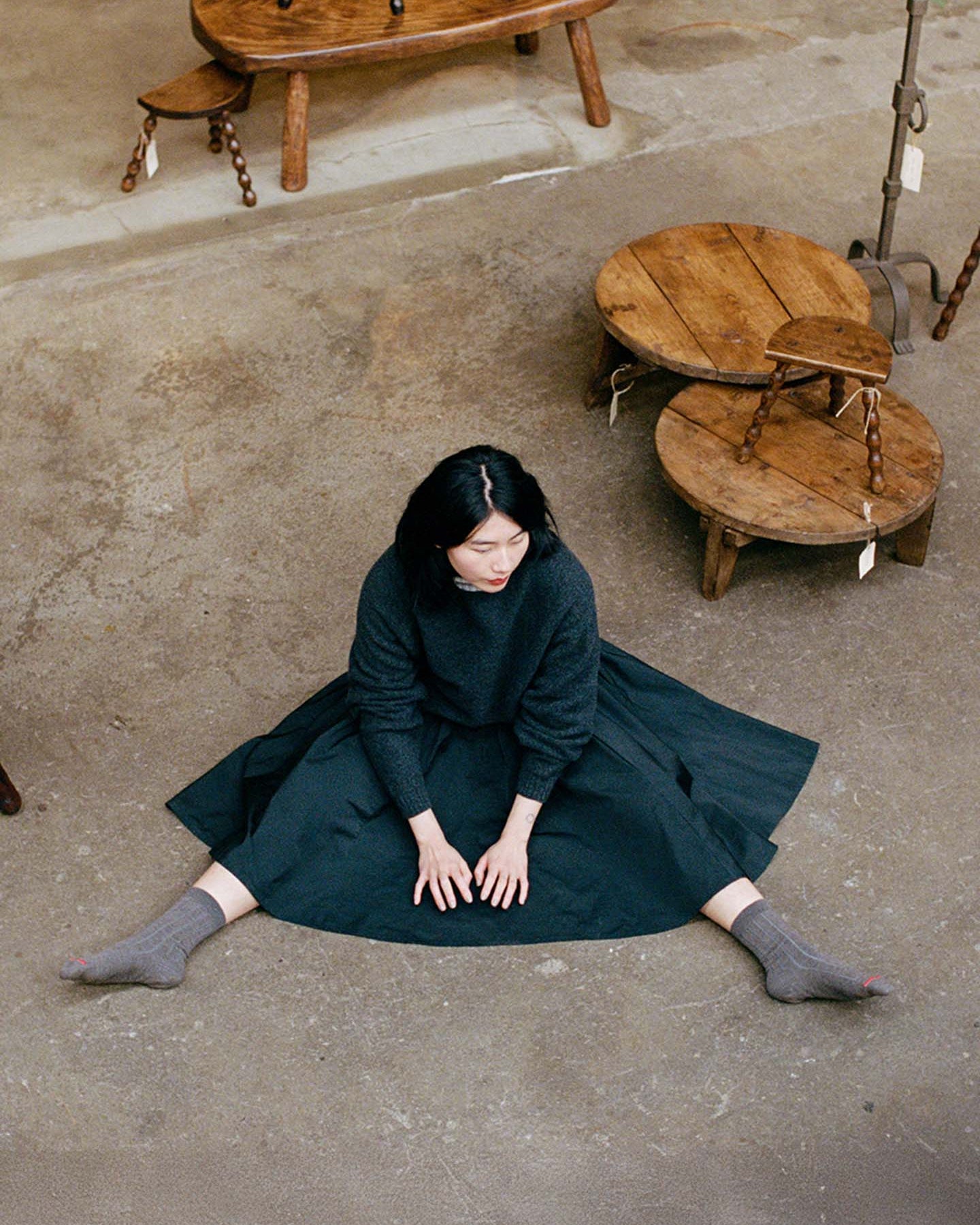 Person stands on a wooden stool, wearing a dark sweater, dark skirt, and Tip Toe Crew Socks – Grey & Cherry Trim, holding part of the skirt. Wooden wall in the background.