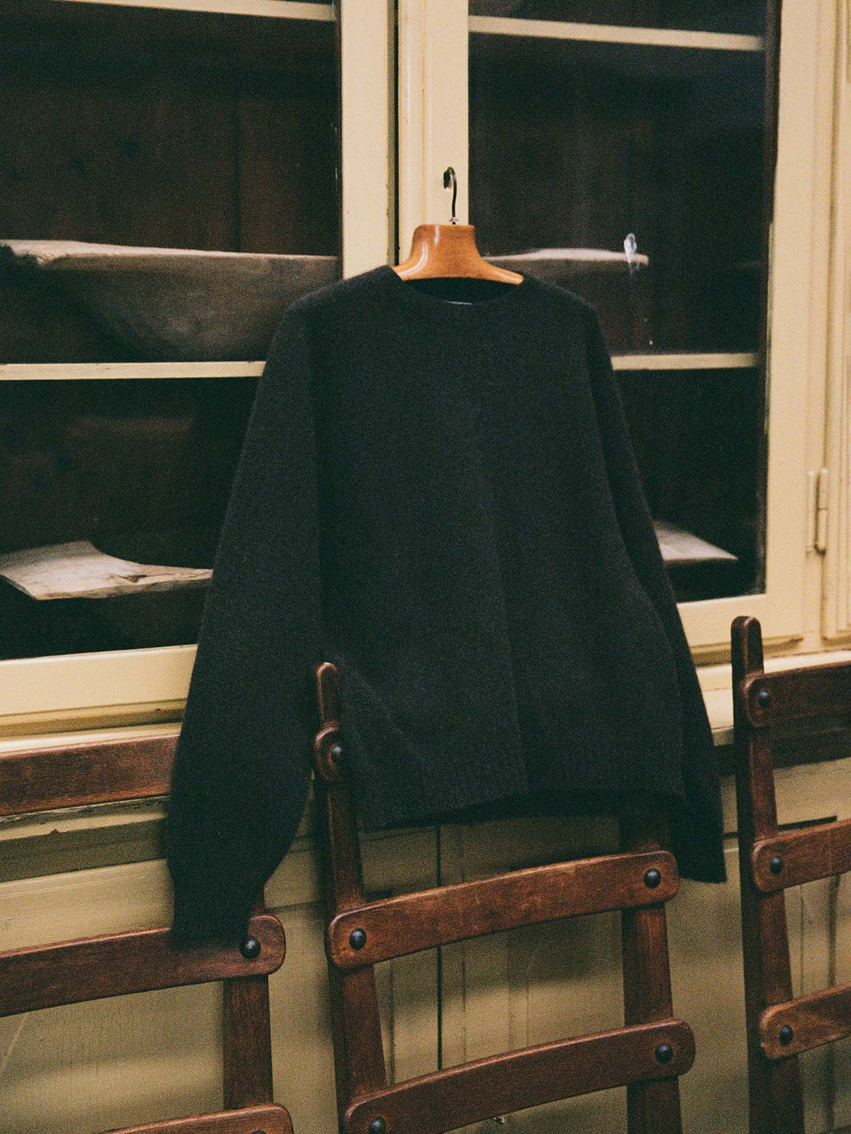 The Poet Knit – Almost Black, a relaxed fit sweater, hangs on a wooden hanger before a glass-front cabinet, with wooden chairs placed below.