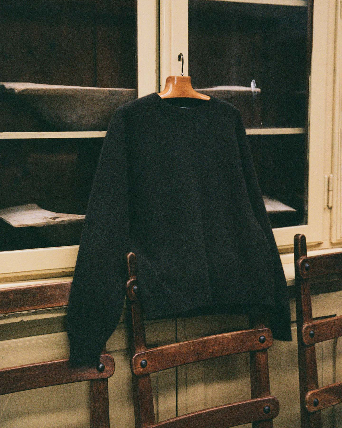 The Poet Knit – Almost Black, a relaxed fit sweater, hangs on a wooden hanger before a glass-front cabinet, with wooden chairs placed below.