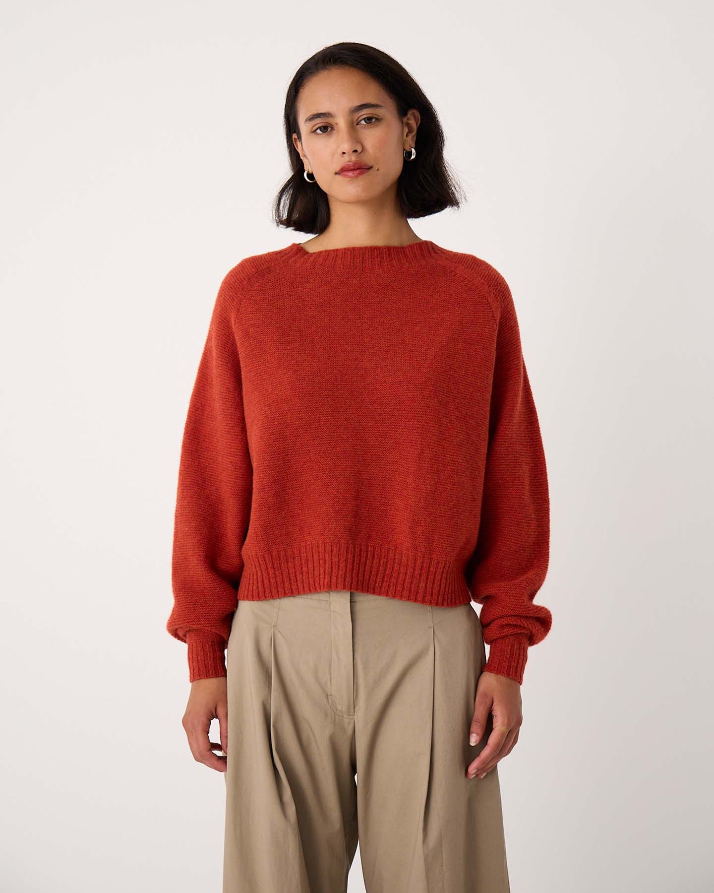 A petite woman stands facing forward, wearing the Poet Knit – Amber in a relaxed fit with beige pants against a plain white background.