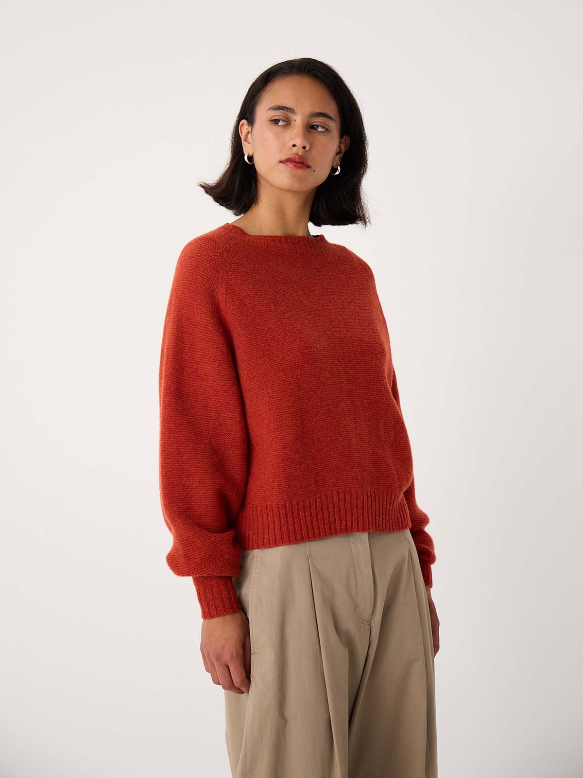 A petite woman wearing the Poet Knit – Amber, a red cropped sweater, and beige pleated pants stands against a plain white background, looking slightly to the side.
