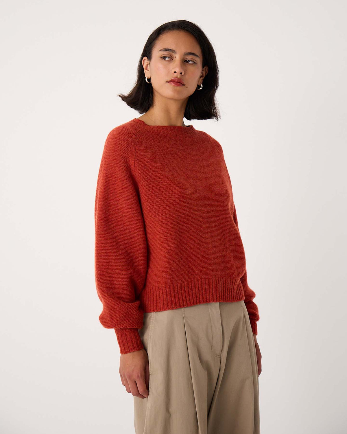 A petite woman wearing the Poet Knit – Amber, a red cropped sweater, and beige pleated pants stands against a plain white background, looking slightly to the side.