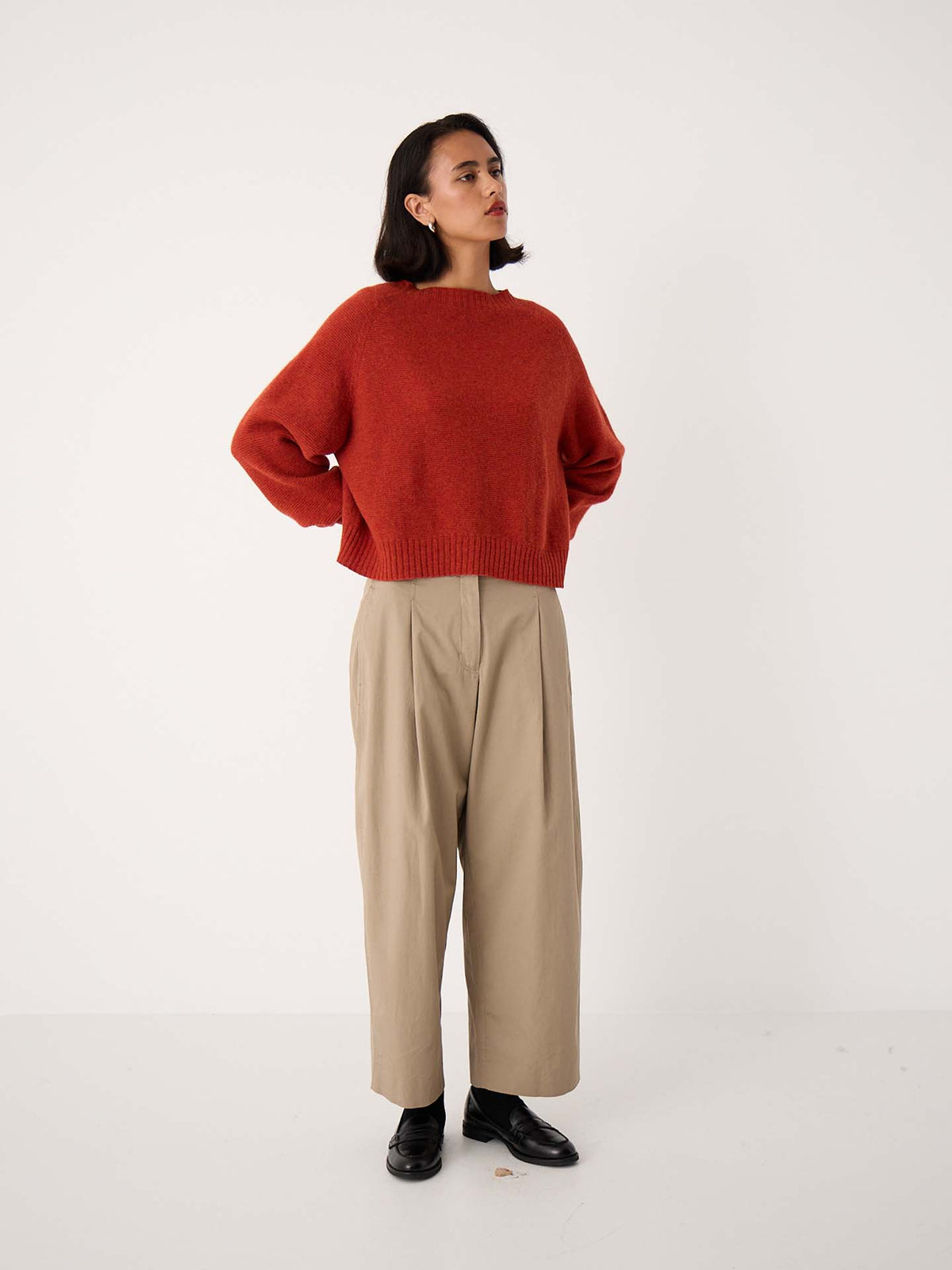 A woman stands against a plain background in the Poet Knit – Amber, a relaxed-fit red sweater, paired with wide-leg beige pants ideal for petite frames and black shoes, hands on hips, gazing to the side.