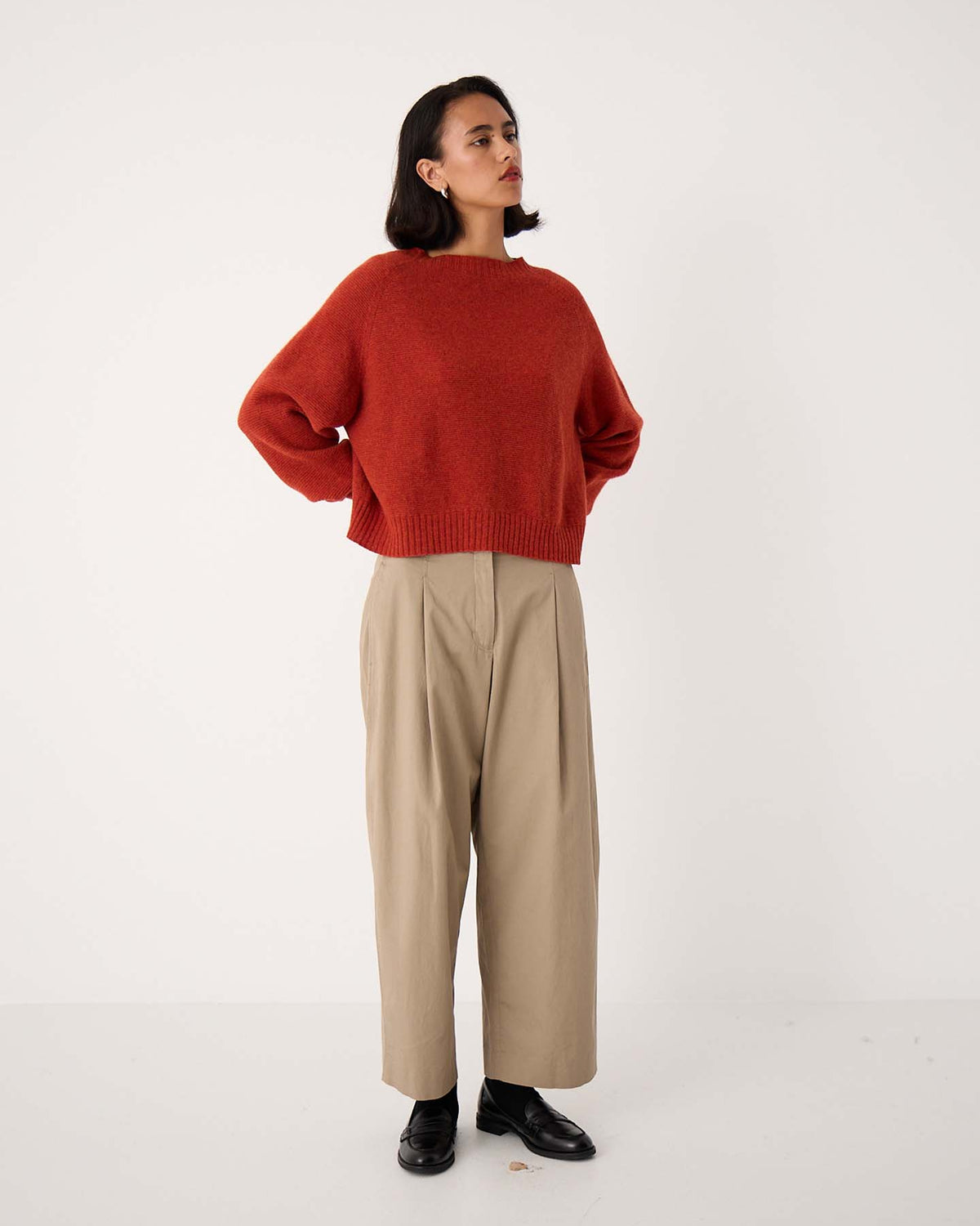 A woman stands against a plain background in the Poet Knit – Amber, a relaxed-fit red sweater, paired with wide-leg beige pants ideal for petite frames and black shoes, hands on hips, gazing to the side.