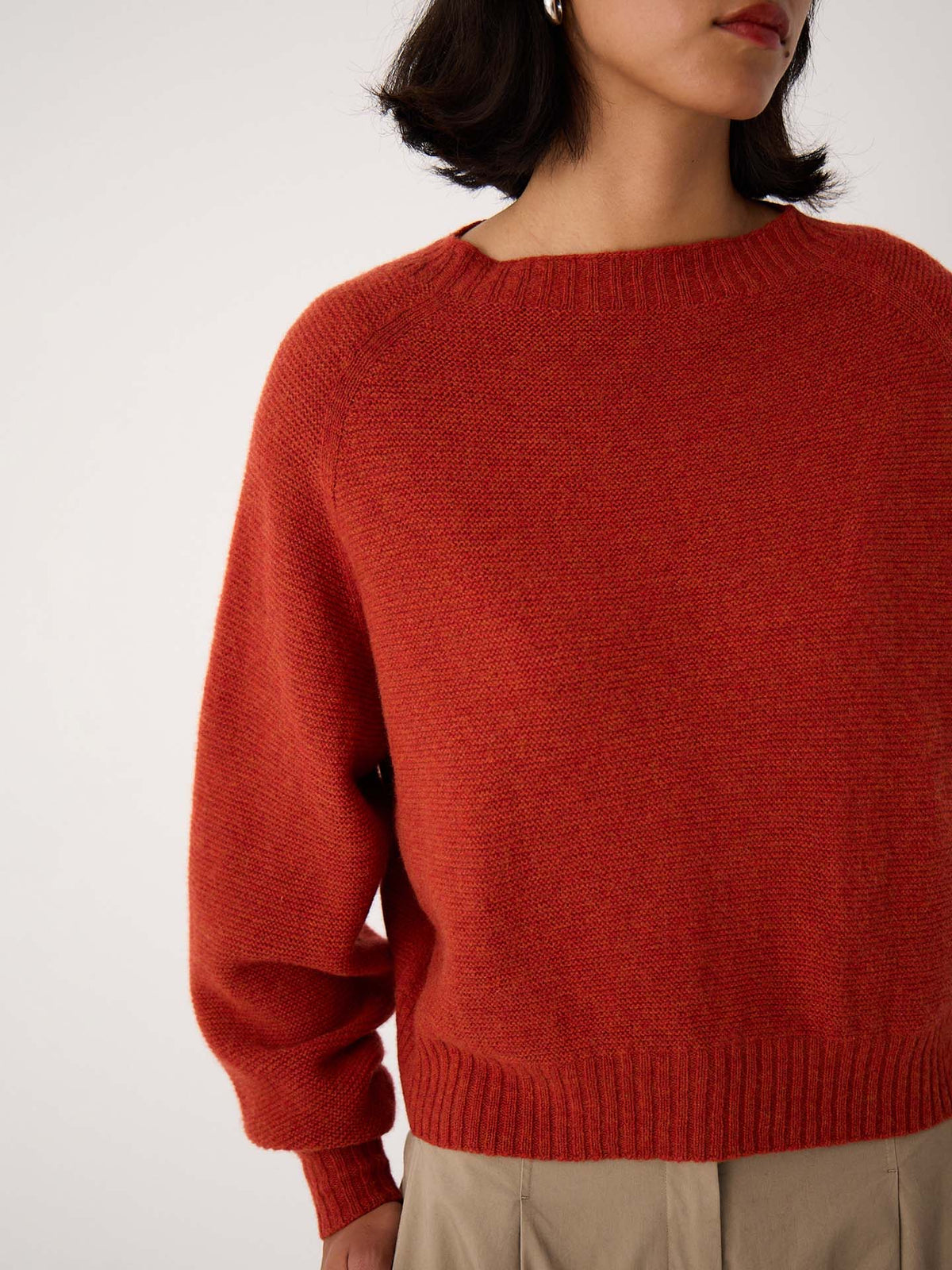 A petite woman with short dark hair models the Poet Knit – Amber, a relaxed-fit red sweater, paired with beige pants. The image is cropped from shoulders to hips and set against a plain background.