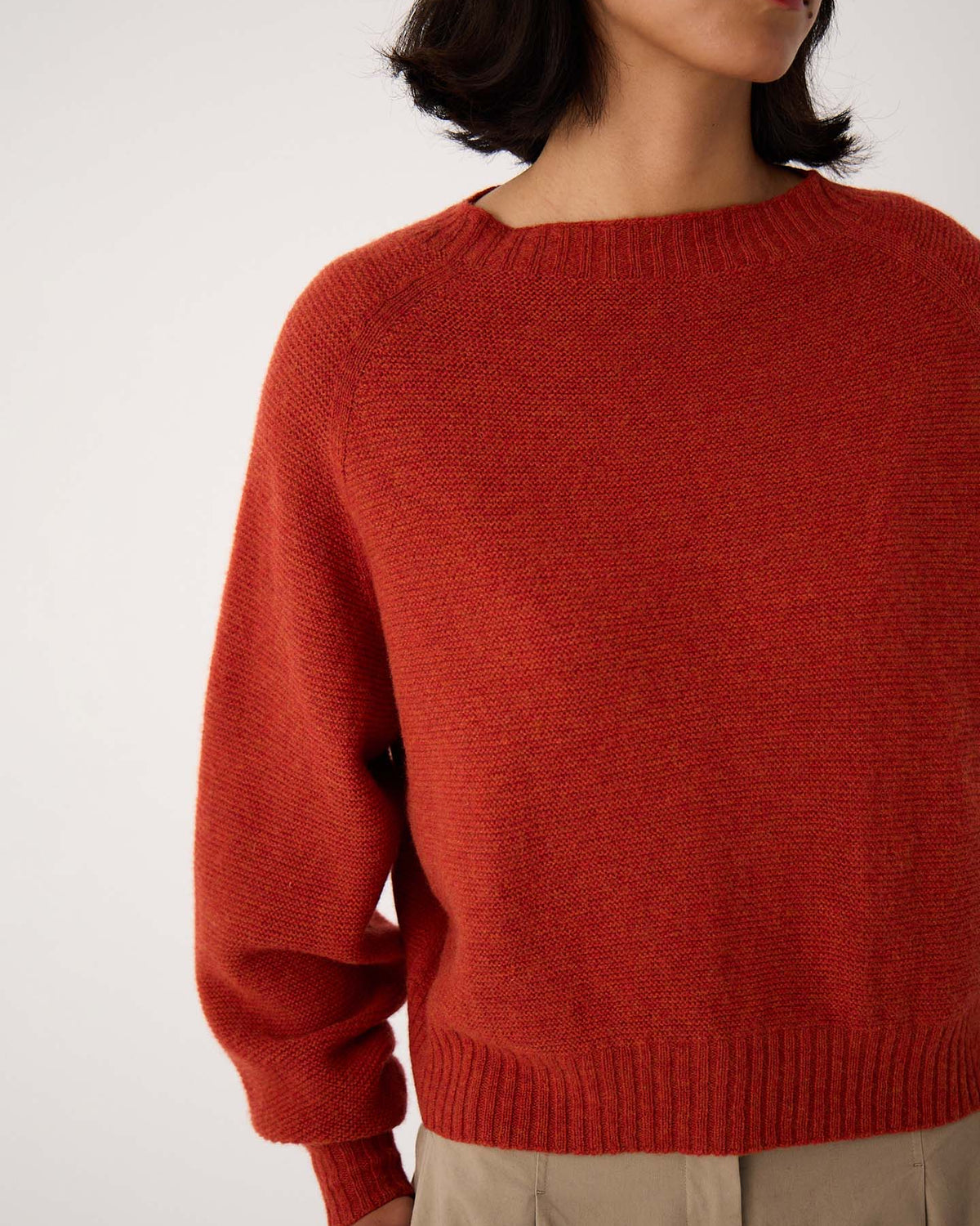 A petite woman with short dark hair models the Poet Knit – Amber, a relaxed-fit red sweater, paired with beige pants. The image is cropped from shoulders to hips and set against a plain background.